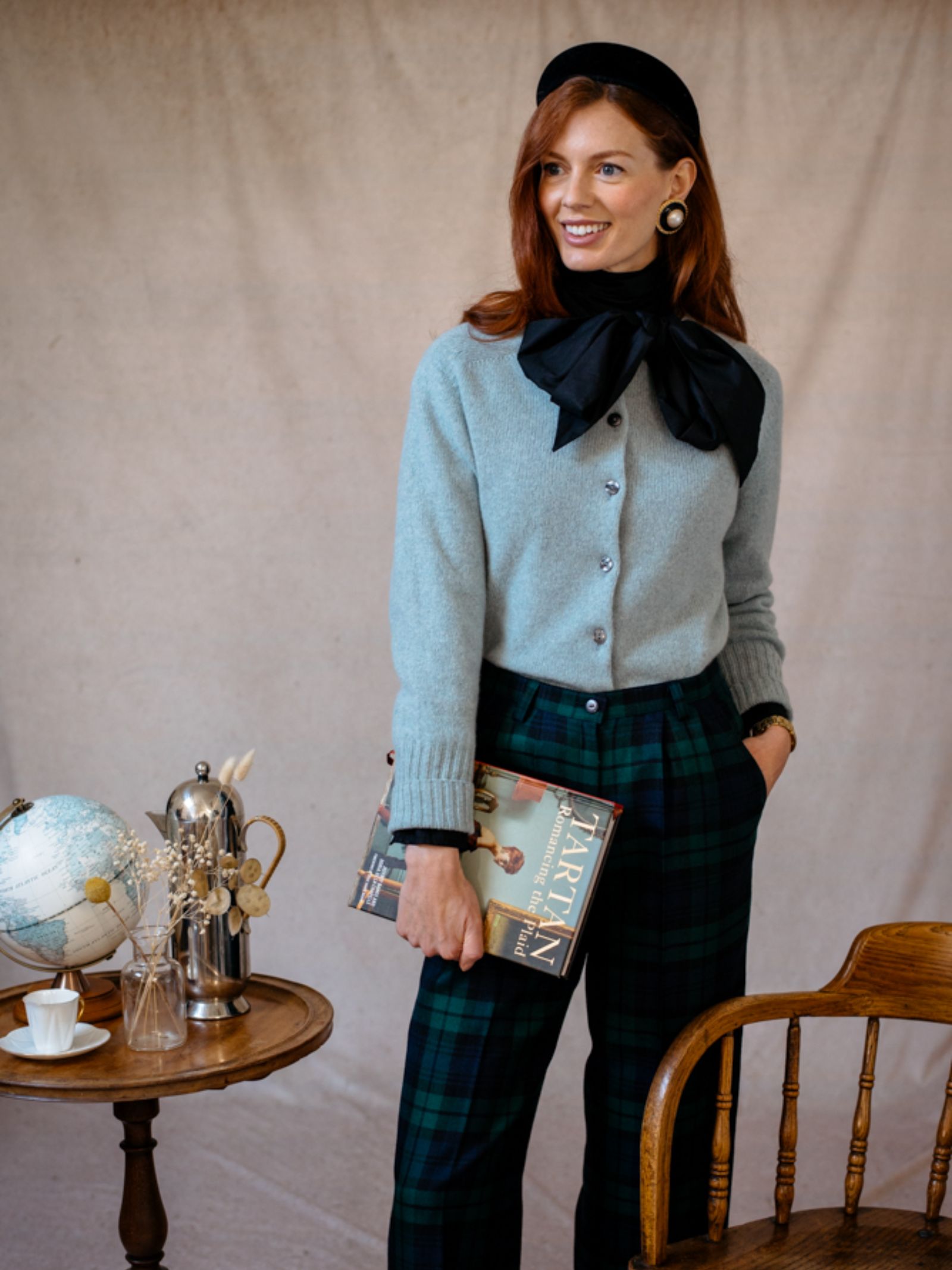 A smiling woman wears a Campbell's of Beauly Geelong Lambswool Crew Cardigan, plaid pants, and a large black bow while holding a book. A globe, teapot, and teacup sit on a small round table beside her against a plain indoor backdrop.
