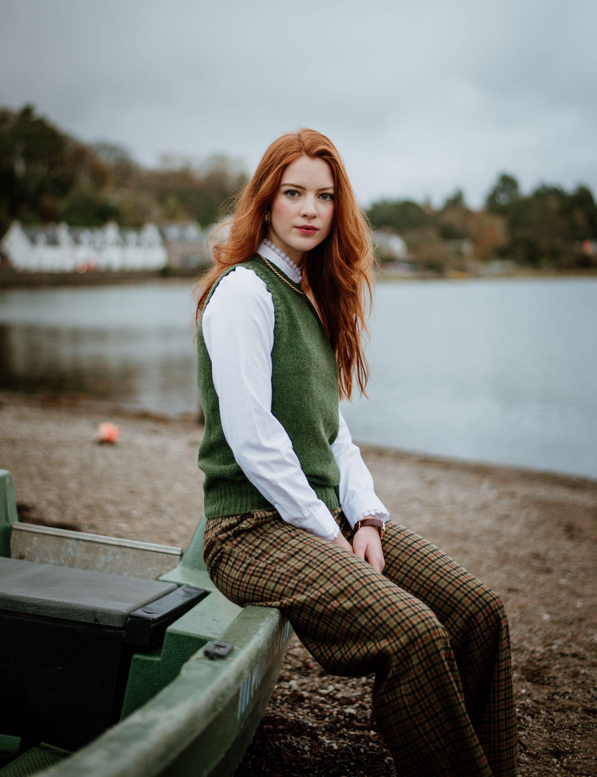 A woman with long red hair sits on the edge of a green boat by a calm lake, wearing a white shirt, green vest, and plaid pants. Trees and houses are visible in the background under a cloudy sky.