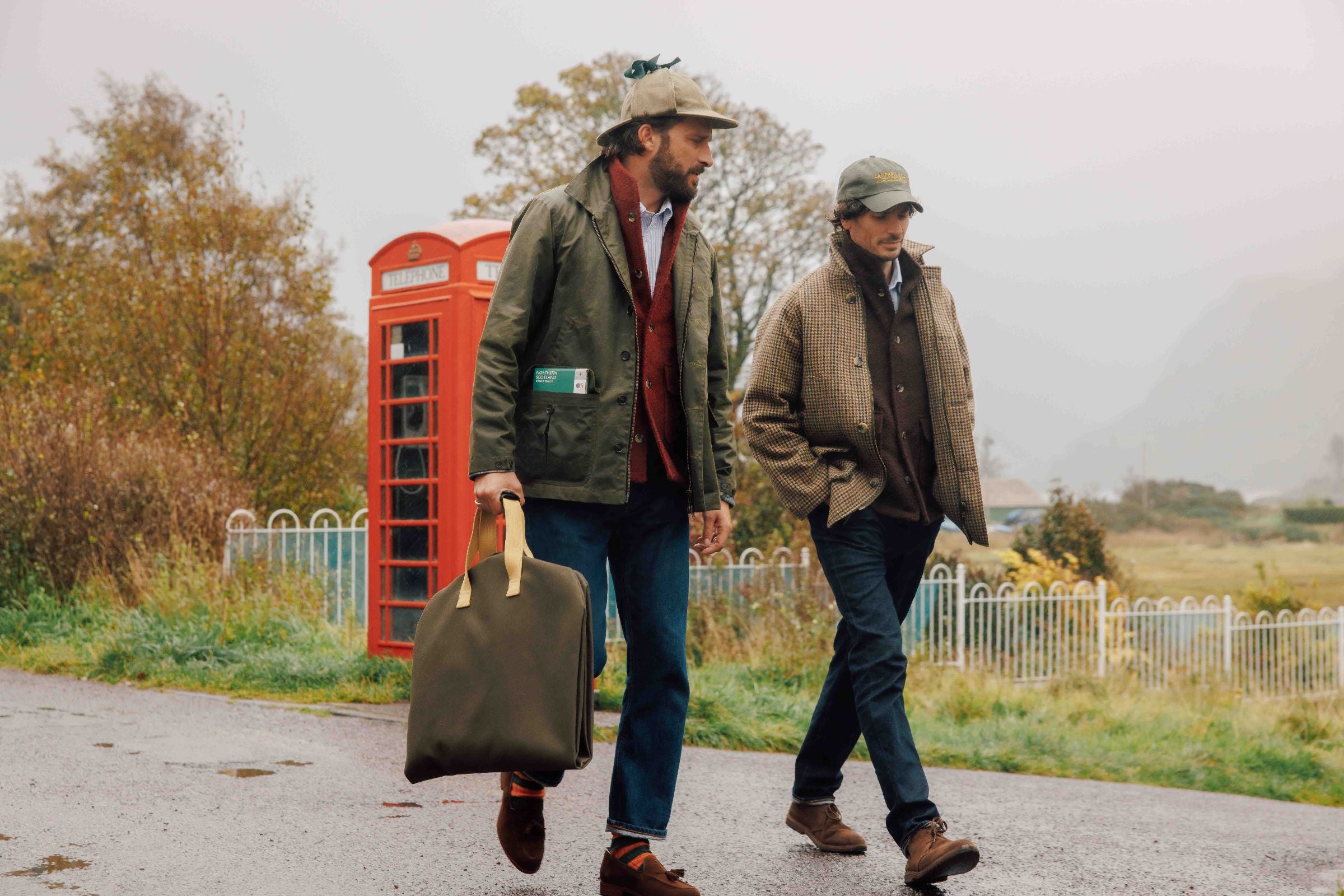 Two men walk on a rural road near a red British phone box. Both wear casual jackets, jeans, and caps; one carries a large green bag. Misty weather and autumn trees are visible in the background.