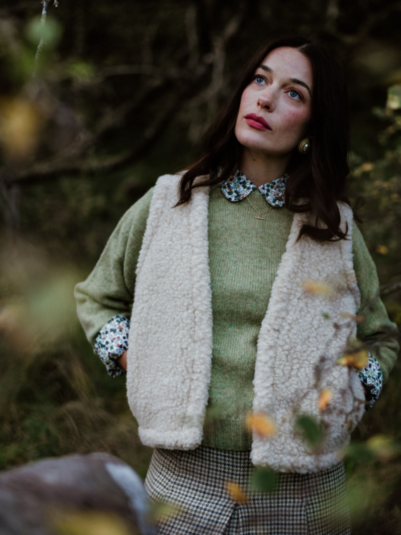 A woman with long brown hair stands outdoors in a Campbell's of Beauly Wool Shrug, its green hue and floral collar layered over a cream sherpa vest and plaid pants—stylish lightweight warmth among blurred trees and foliage.