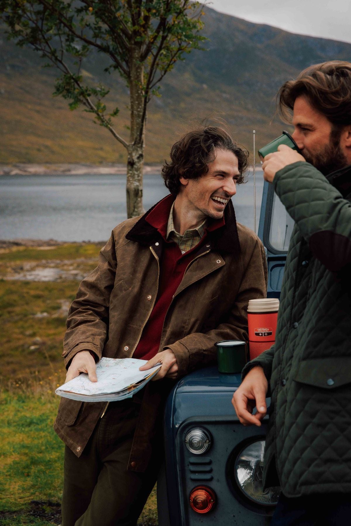 Two men stand by a blue vehicle near a lake and hills. One man, smiling and holding a map, leans on the car, while the other drinks from a mug. They wear outdoor jackets, suggesting a road trip or adventure.