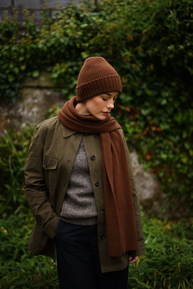 A person wearing a brown knit hat and scarf, an olive green jacket, and a gray sweater stands outdoors in front of lush greenery, looking down with hands in their pockets.