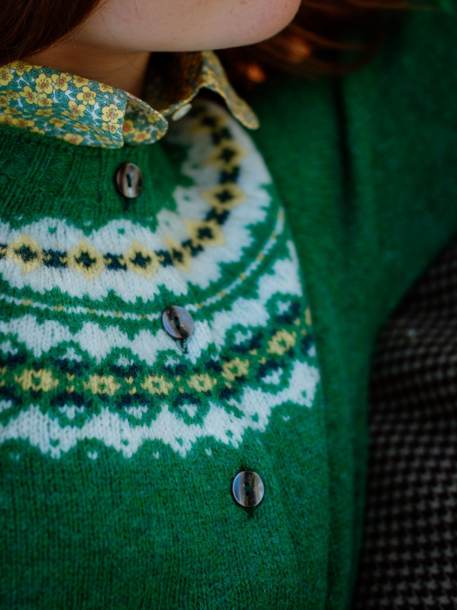 Close-up of a person in the Campbell's of Beauly Auld Stock Shetland Fairisle Cardigan, crafted from pure new wool in green with a white and yellow Nordic pattern, layered over a floral-collared shirt.