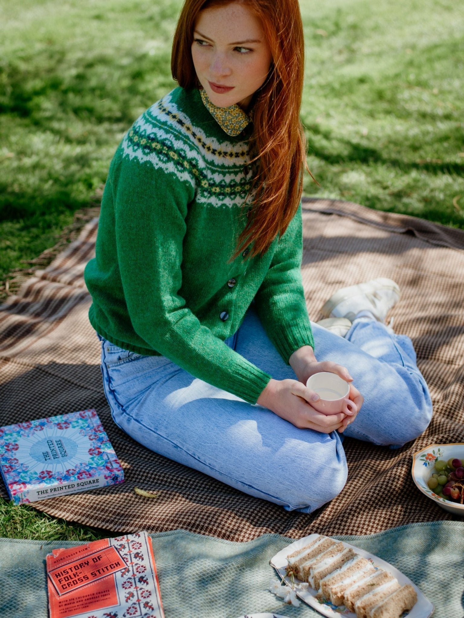 A woman with long red hair in a Campbell's of Beauly Auld Stock Shetland Fairisle Cardigan and light jeans sits on a picnic blanket, holding a cup. Beside her are books, sliced cake, and grapes on the grass.