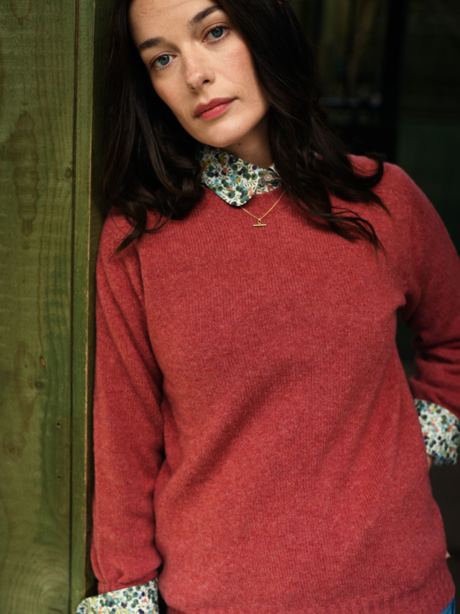 A woman with long dark hair leans against a green wooden wall, wearing a red Campbell's of Beauly Geelong Lambswool Crew Neck Jumper over a floral-collared shirt, gazing at the camera with a neutral expression.