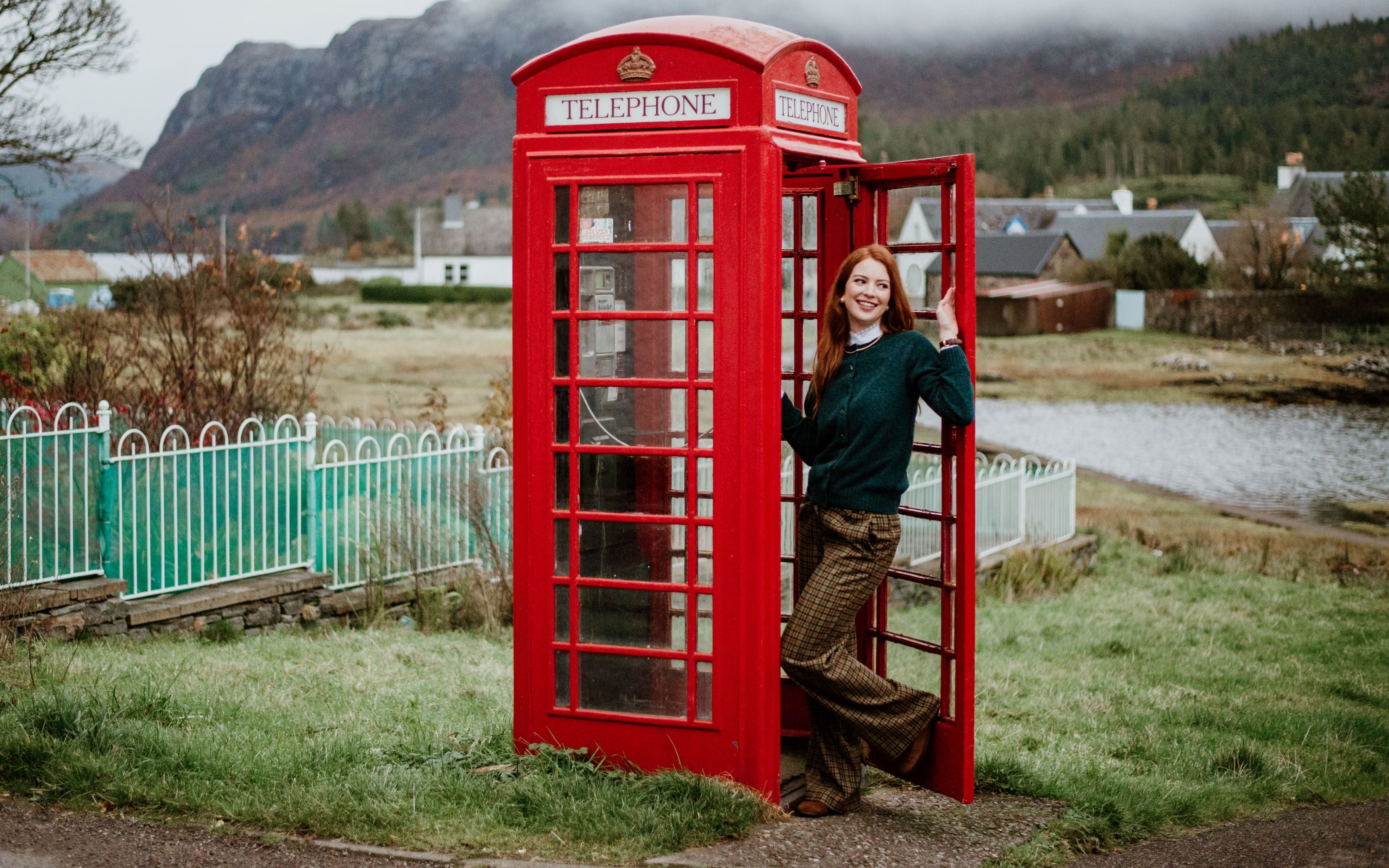 A smiling woman stands in the doorway of a classic red British phone booth, surrounded by grass, a pond, houses, and misty hills in the background.