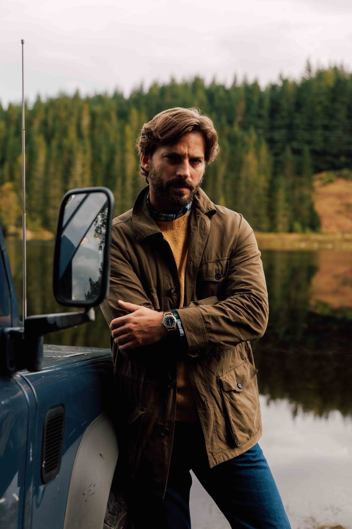 A man with wavy hair and a beard stands by a blue vehicle near a calm lake, wearing a brown jacket, mustard sweater, and watch, with a background of dense evergreen trees.