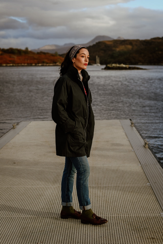 A woman wearing a dark coat, jeans, and boots stands on a dock by a body of water, with hills and cloudy skies in the background. She gazes into the distance with her hands in her coat pockets.