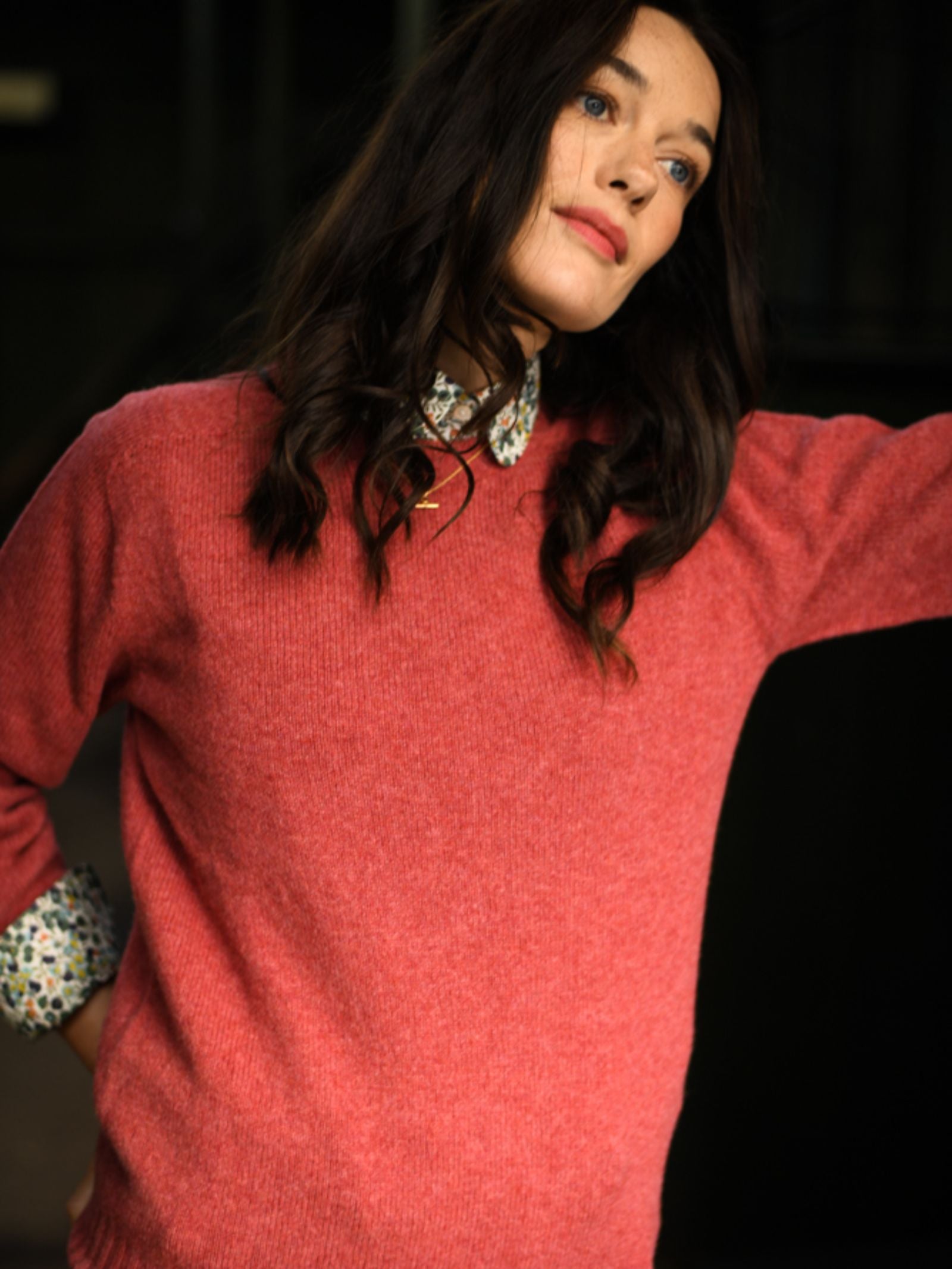 A woman with wavy dark hair and blue eyes wears the pink Geelong Lambswool Crew Neck Jumper by Campbell's of Beauly over a floral-collared shirt, standing with one arm raised against a dark, blurred background.