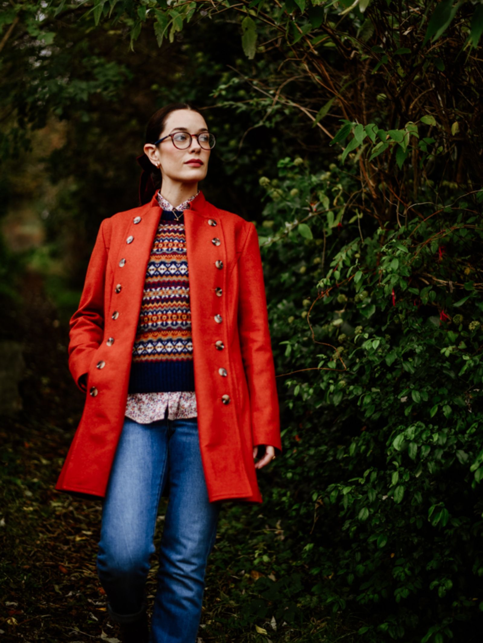 Wearing a vibrant red Pirate Coat by Campbell's of Beauly, a person in a patterned sweater, blue jeans, and glasses strolls outdoors along a leafy path bordered by lush green foliage.