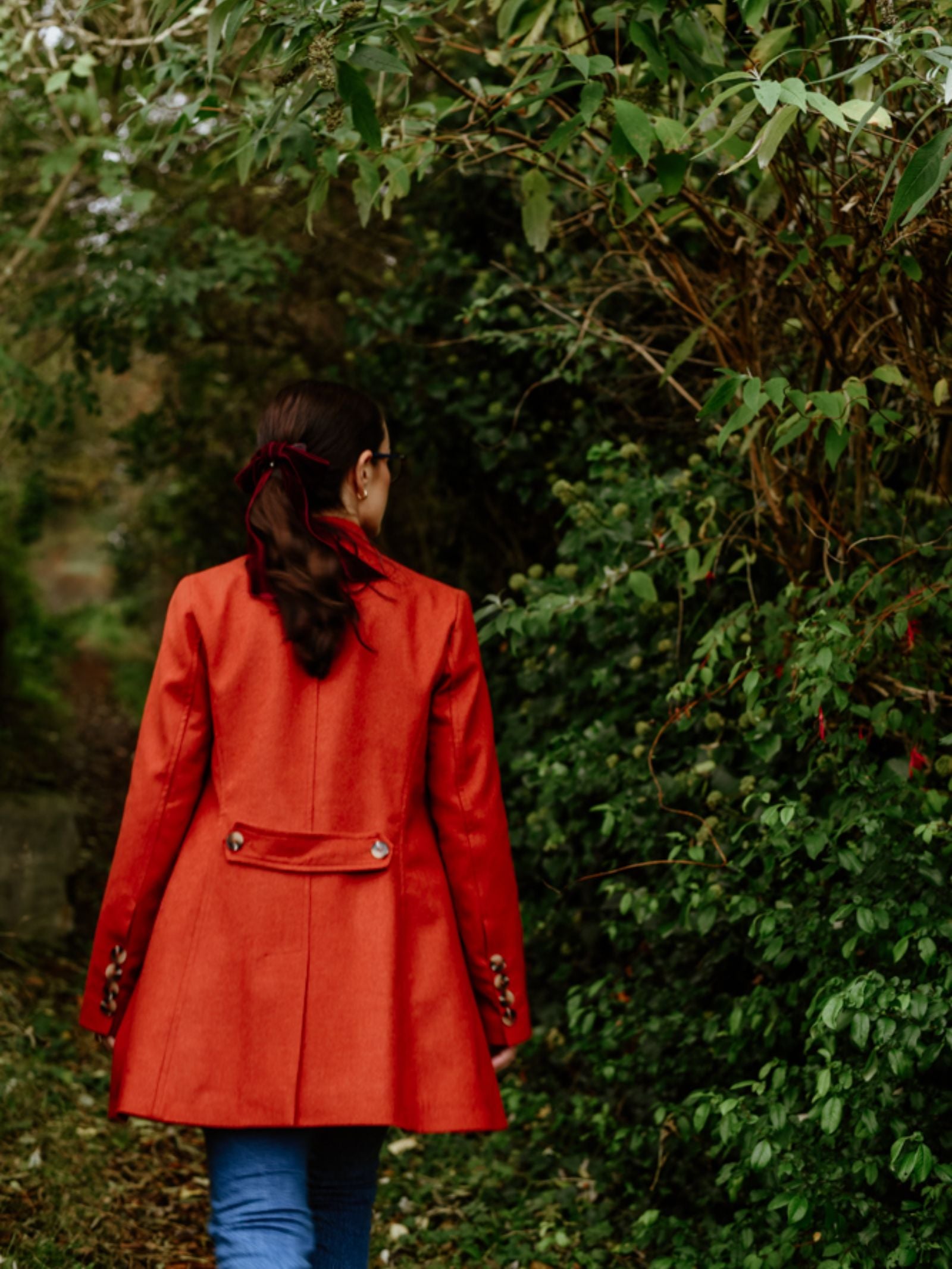 A person with long brown hair tied with a ribbon walks away on a path lined with greenery, wearing blue jeans and a bright red Pirate Coat by Campbell's of Beauly.