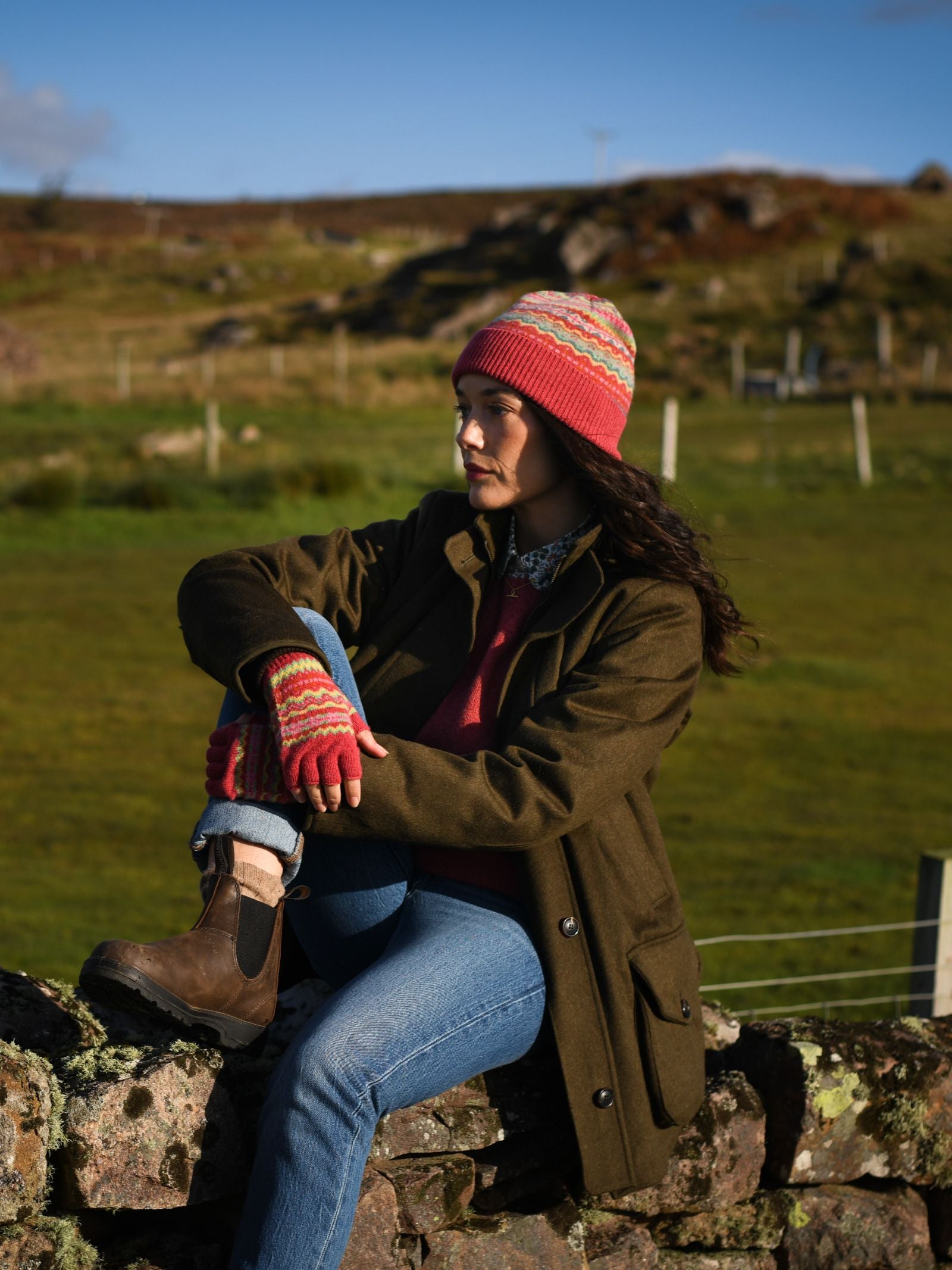 A woman in the Auld Stock Lambswool Fairisle Hat by Campbell's of Beauly sits on a stone wall, her vibrant winter look—hat, gloves, green jacket, blue jeans, and boots—standing out against a grassy rural landscape under a clear blue sky.