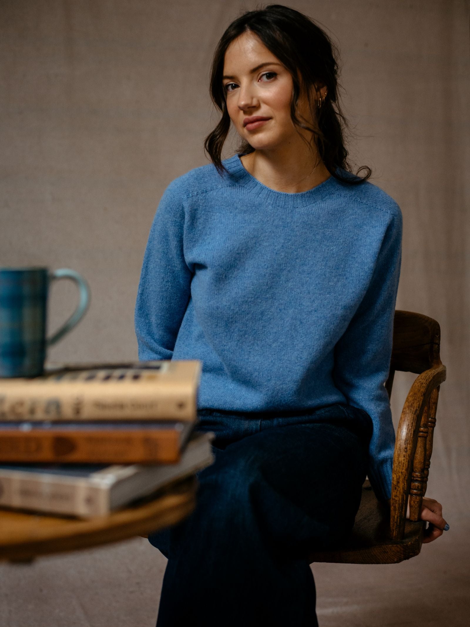 A dark-haired woman wears a blue Auld Stock Geelong Lambswool Crew Neck Jumper by Campbell's of Beauly, sitting on a wooden chair and looking at the camera. In front, books and a blue checkered mug sit on the table against a simple backdrop.