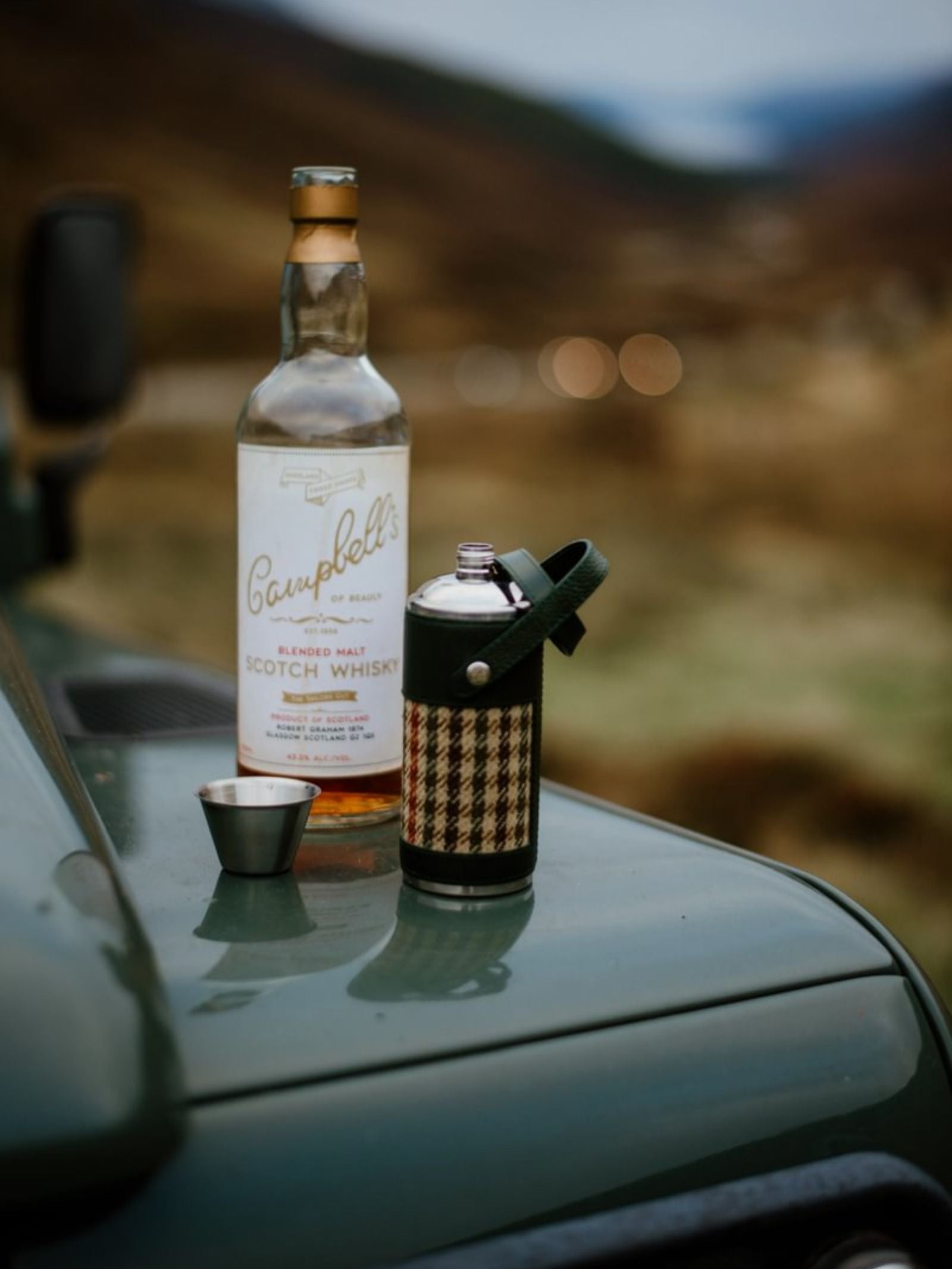 An 8oz Forest Green Hunter Flask by Campbell's of Beauly x Ettinger, a bottle of Campbell’s Scotch whisky, and a metal cup rest on the hood of a green vehicle with a blurred scenic landscape in the background.