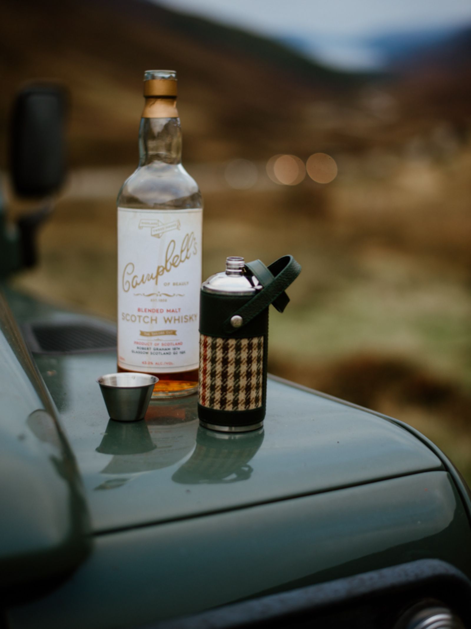 An 8oz Bordeaux Hunter Flask by Campbell's of Beauly x Ettinger, a metal cup, and a bottle of Scotch whisky rest on the hood of a green vehicle against a blurred outdoor backdrop.