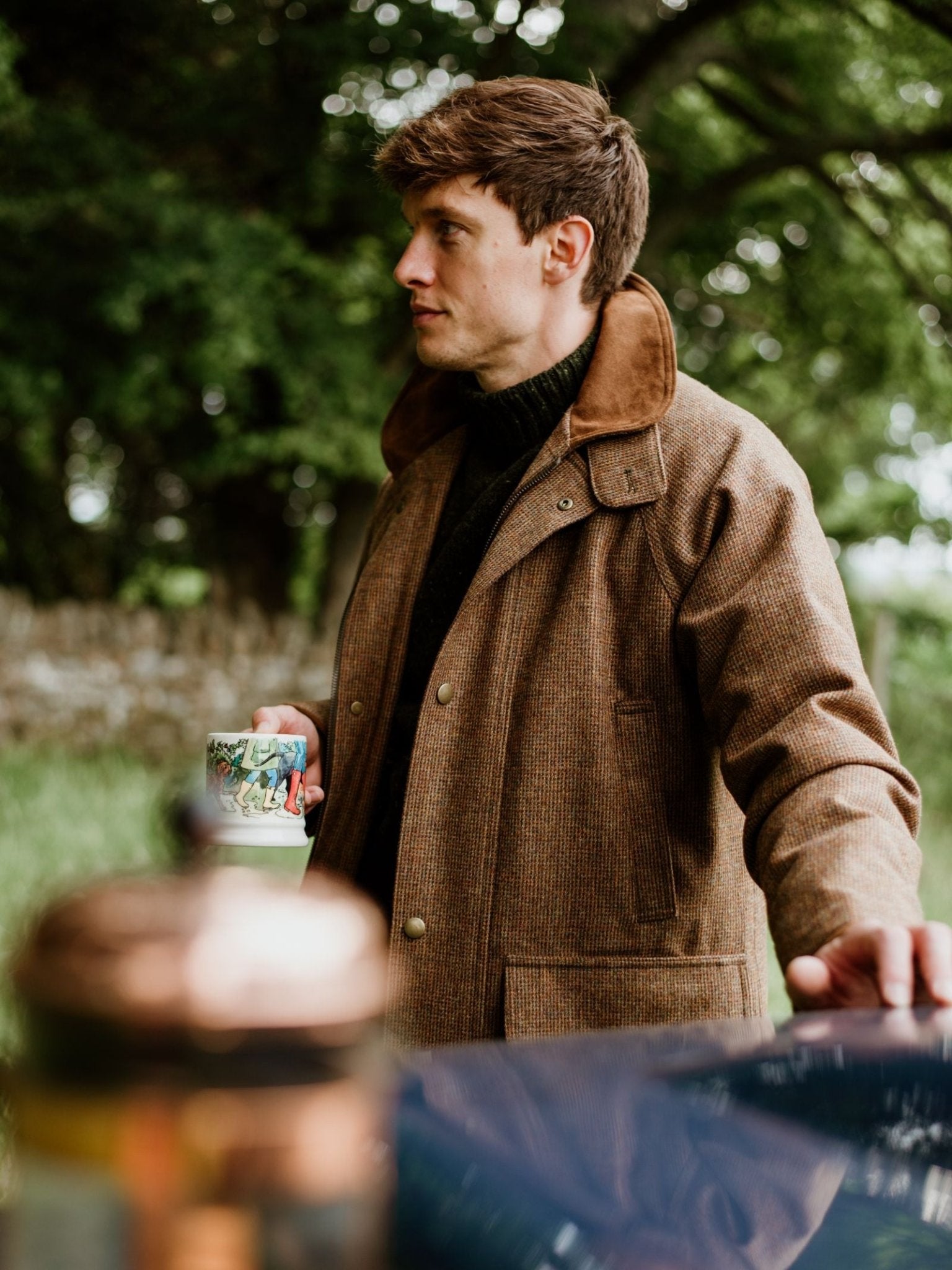 A man in a Campbell's of Beauly Tweed Field Coat stands outdoors by a table, holding a colorful mug with trees and greenery in the background.