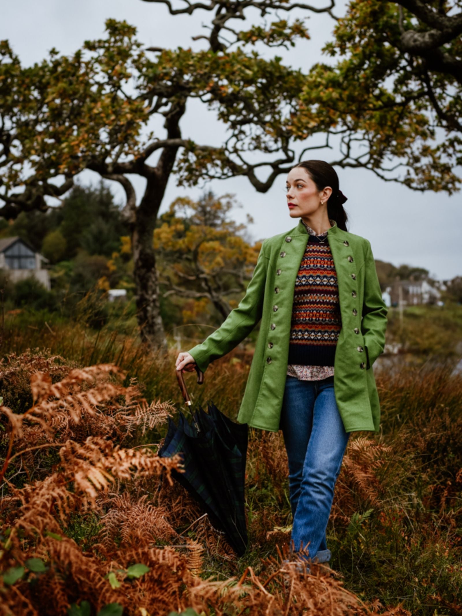 A woman wearing a green Pirate Coat by Campbell's of Beauly and a patterned sweater stands outdoors among autumn ferns, holding a closed umbrella. Yellowing trees and distant houses appear under a cloudy sky behind her.