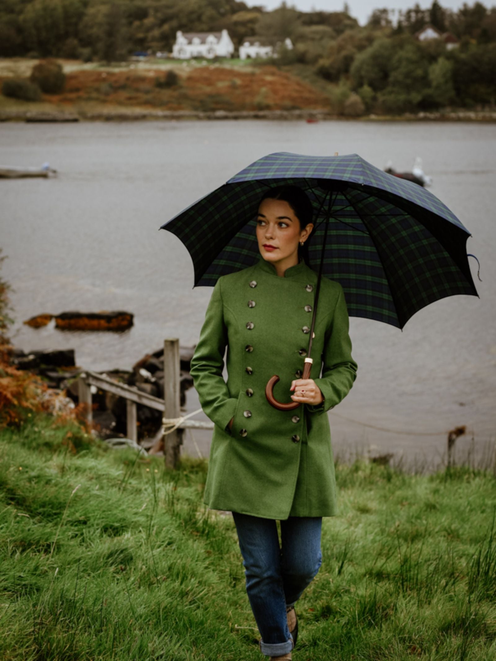 A woman wears a green Pirate Coat by Campbell's of Beauly and jeans, holding a plaid umbrella on grass near water, with trees and houses in the background under cloudy skies.