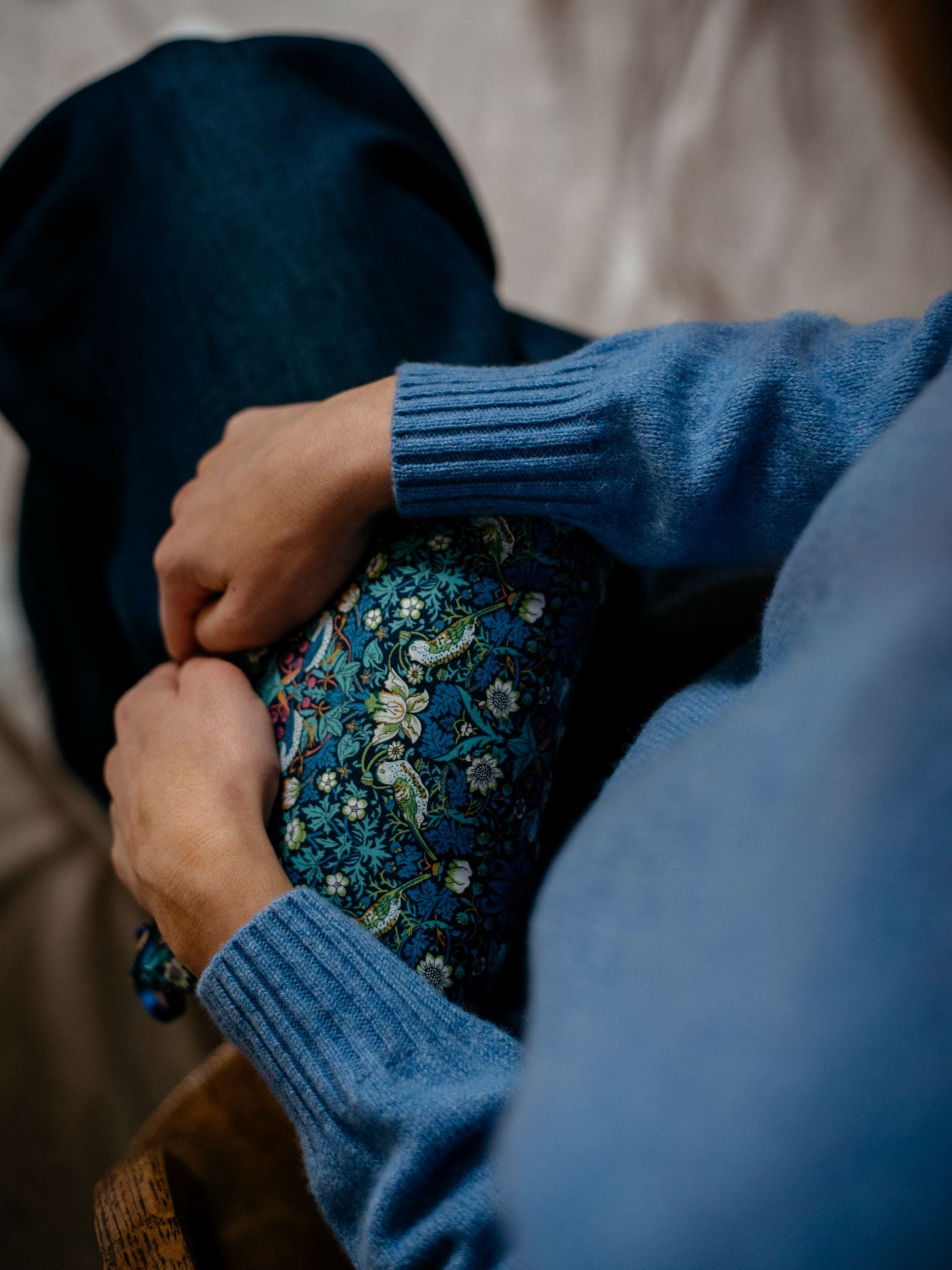 Wearing a Campbell's of Beauly Auld Stock Geelong Lambswool Crew Neck Jumper and jeans, a person sits calmly with hands clasped around their knee, which is draped in blue floral fabric, evoking an introspective mood.