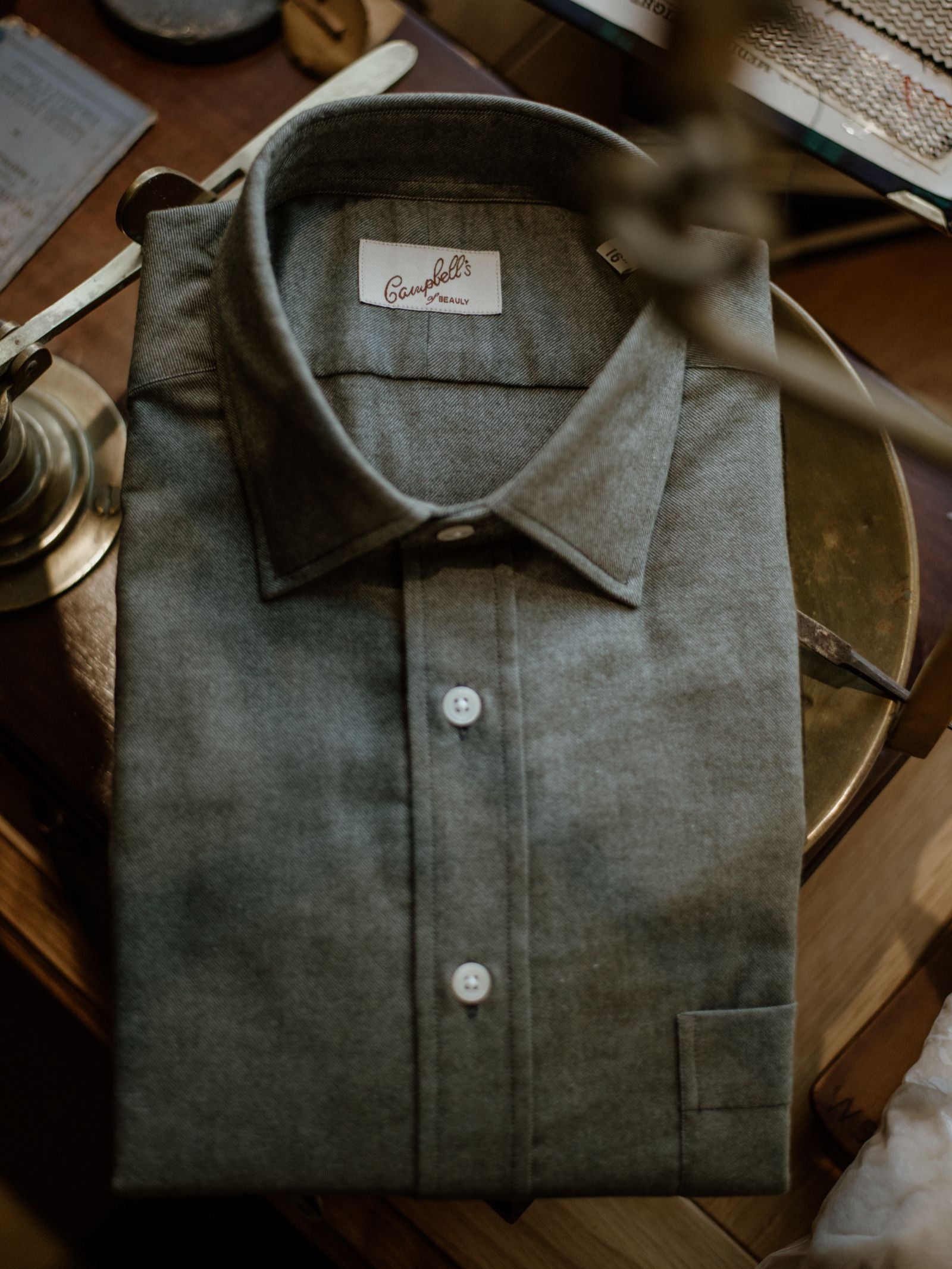 A neatly folded gray Twill Shirt from Campbell's of Beauly rests on a wooden surface beside a brass lamp and other desk accessories.