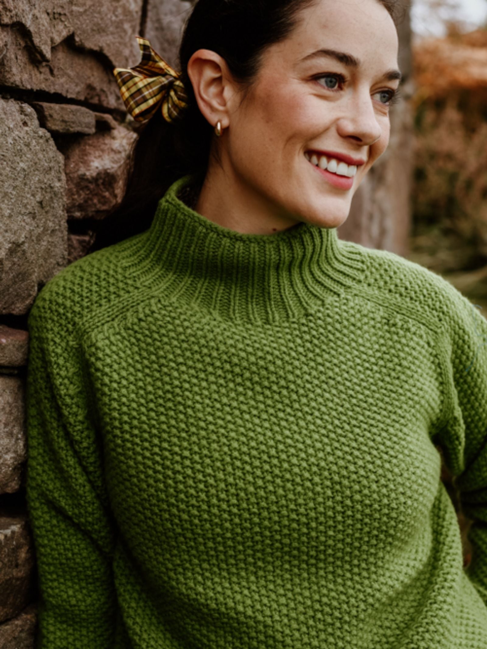 A woman with dark hair tied back in a plaid scrunchie smiles as she leans against a stone wall, wearing the Chunky Moss Stitch Polo by Campbell's of Beauly.