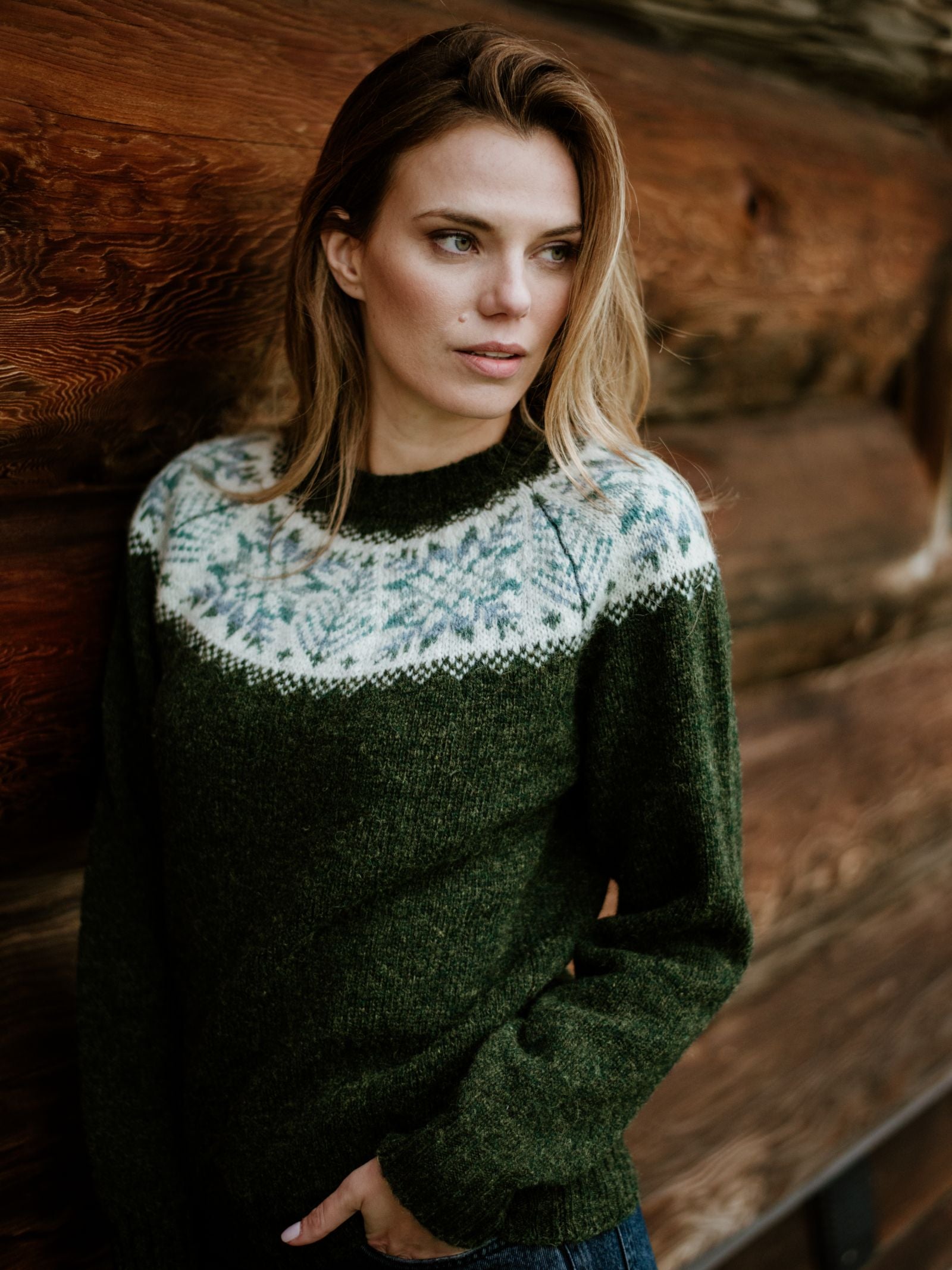 A woman with light brown hair stands by a rustic wooden wall, wearing Campbell's of Beauly Auld Stock Shetland Wool Snowflake Crew Jumper in dark green, featuring a white and blue Fairisle design across the chest and shoulders.
