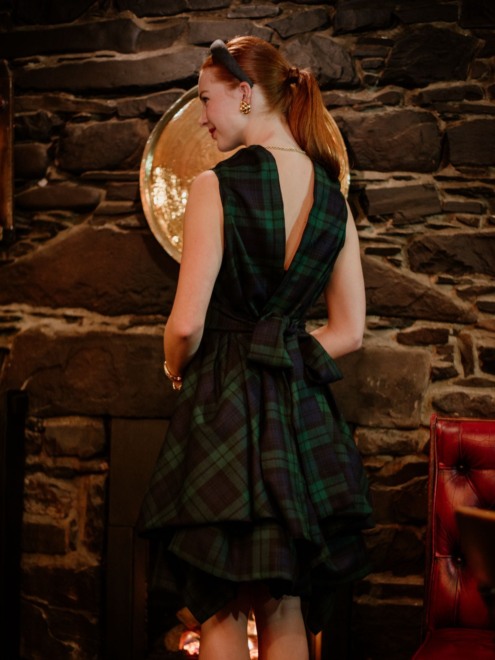A woman with red hair in a ponytail, wearing a Campbell's of Beauly Reversible Wool Wrap Dress with a large back bow, stands facing away from the camera in front of a stone fireplace.