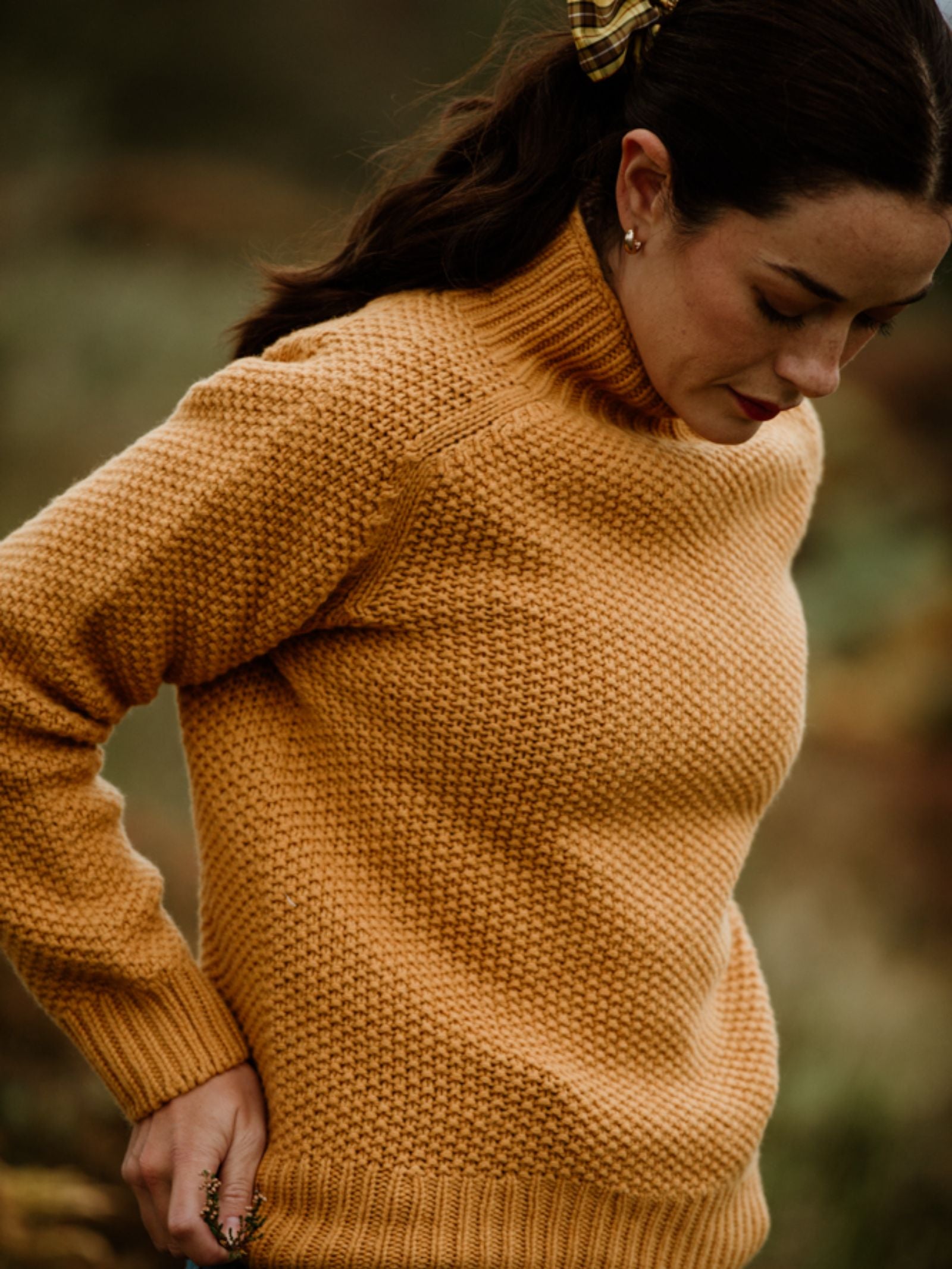 A woman wears the Campbell's of Beauly Auld Stock Chunky Moss Stitch Polo Jumper. Her dark hair is pulled back with a checkered scrunchie as she looks down.