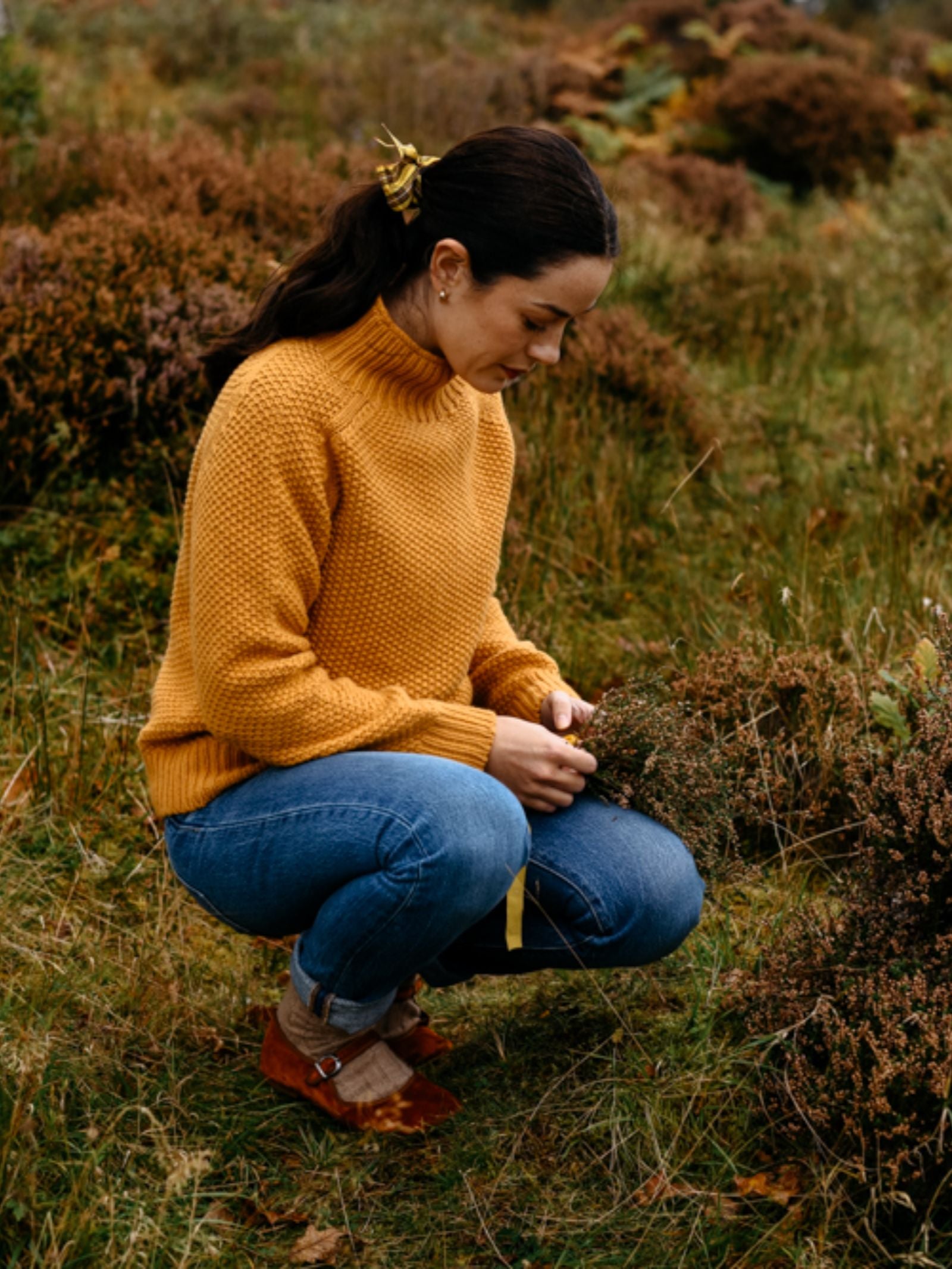 A woman wears the Auld Stock Chunky Moss Stitch Polo Jumper by Campbell's of Beauly with blue jeans, brown shoes, and a yellow hair clip as she crouches in a grassy field examining plants. Green grass and brown foliage surround her.