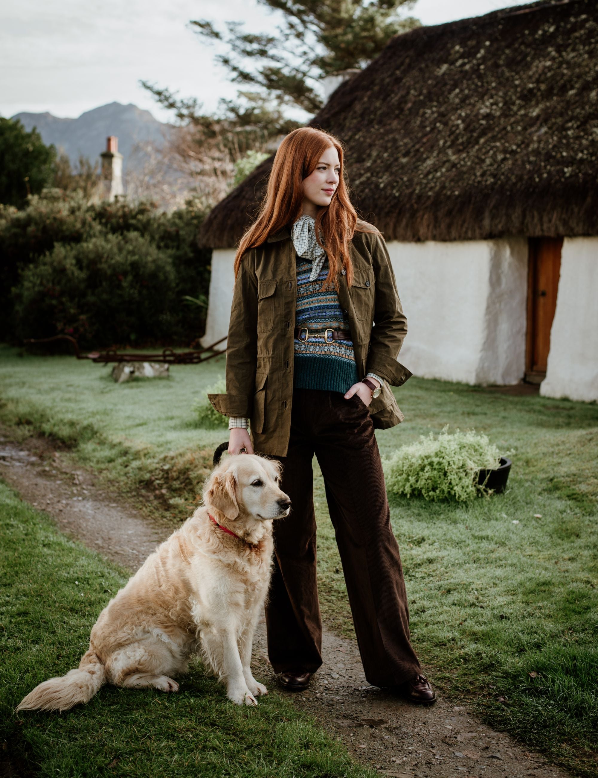 A woman with long red hair stands outdoors beside a golden retriever. She wears a sweater, green jacket, brown pants, and scarf. Behind them is a white cottage with a thatched roof and garden greenery.