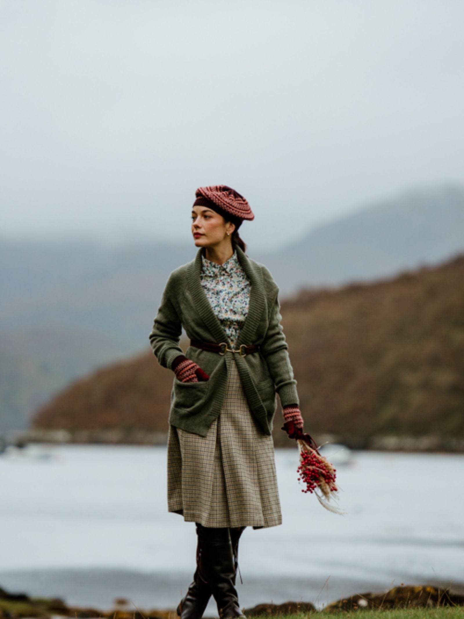 A woman dressed in vintage-inspired attire stands beside a lake, holding a bouquet of flowers. She wears the Fairisle Beret by Campbell’s of Beauly, beautifully crafted from soft lambswool, along with a green cardigan, floral blouse, and plaid skirt. Misty hills in the background evoke the timeless charm of Scottish knitting traditions.