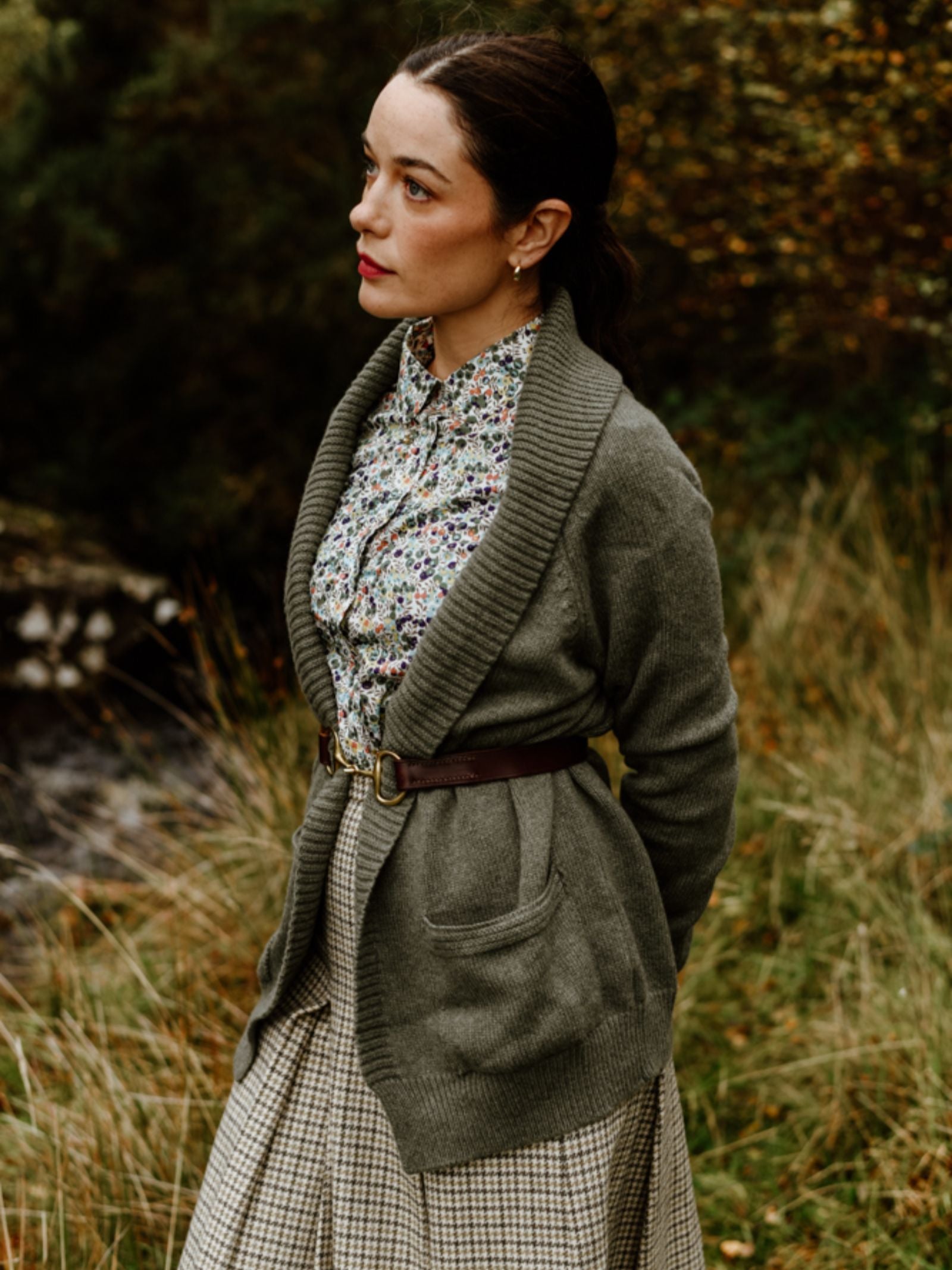 A woman stands outdoors in tall grass wearing a floral blouse, plaid skirt, and a belted Cashmere Shawl Cardigan by Campbell’s of Beauly. Her dark hair is pulled back as she gazes thoughtfully amidst autumn foliage.