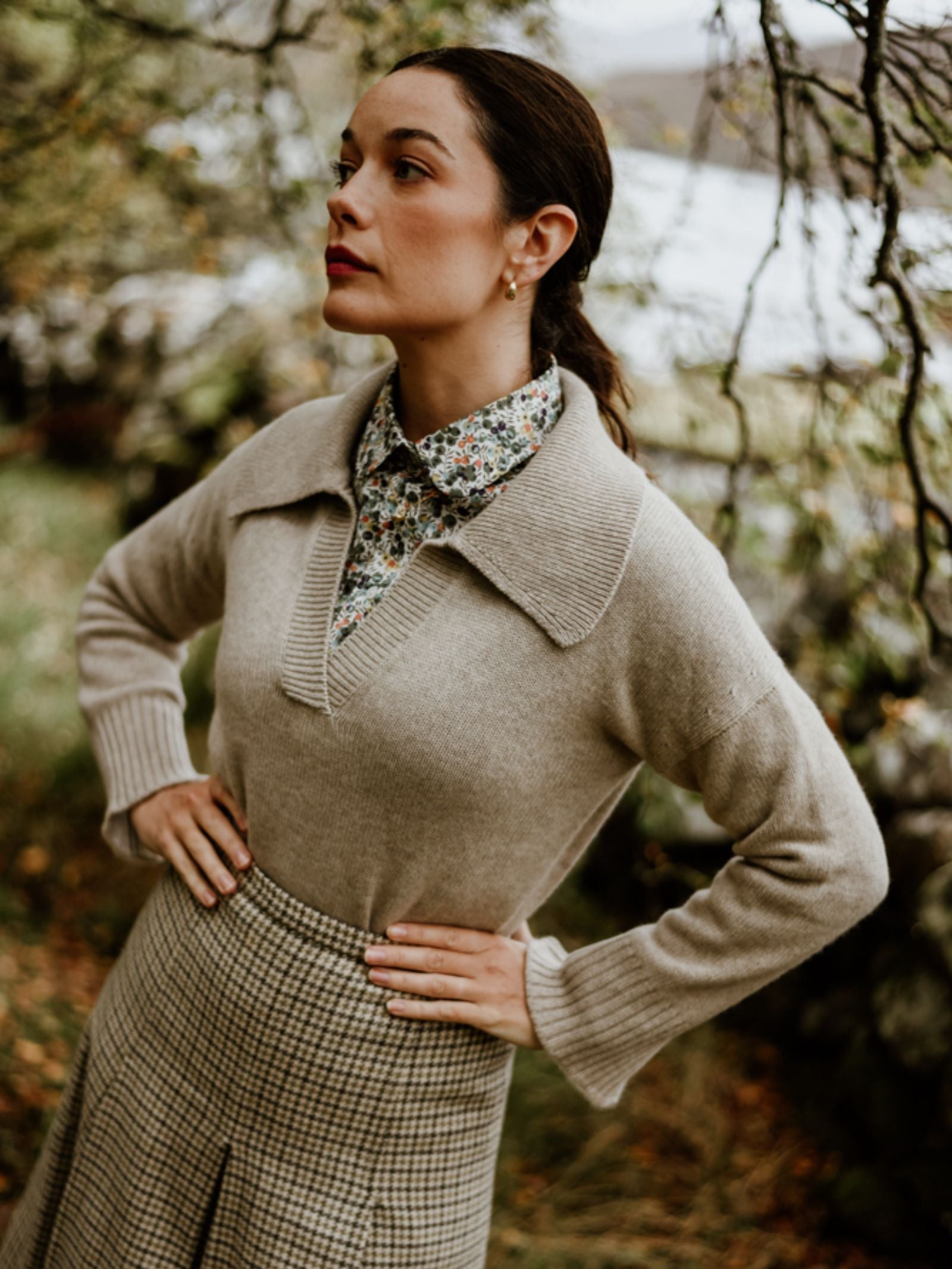 A woman stands outdoors in the Scottish Borders, wearing the Campbell's of Beauly Auld Stock Cashmere Shirt Jumper over a floral collared shirt and plaid skirt, surrounded by greenery with a blurred lake in the background.