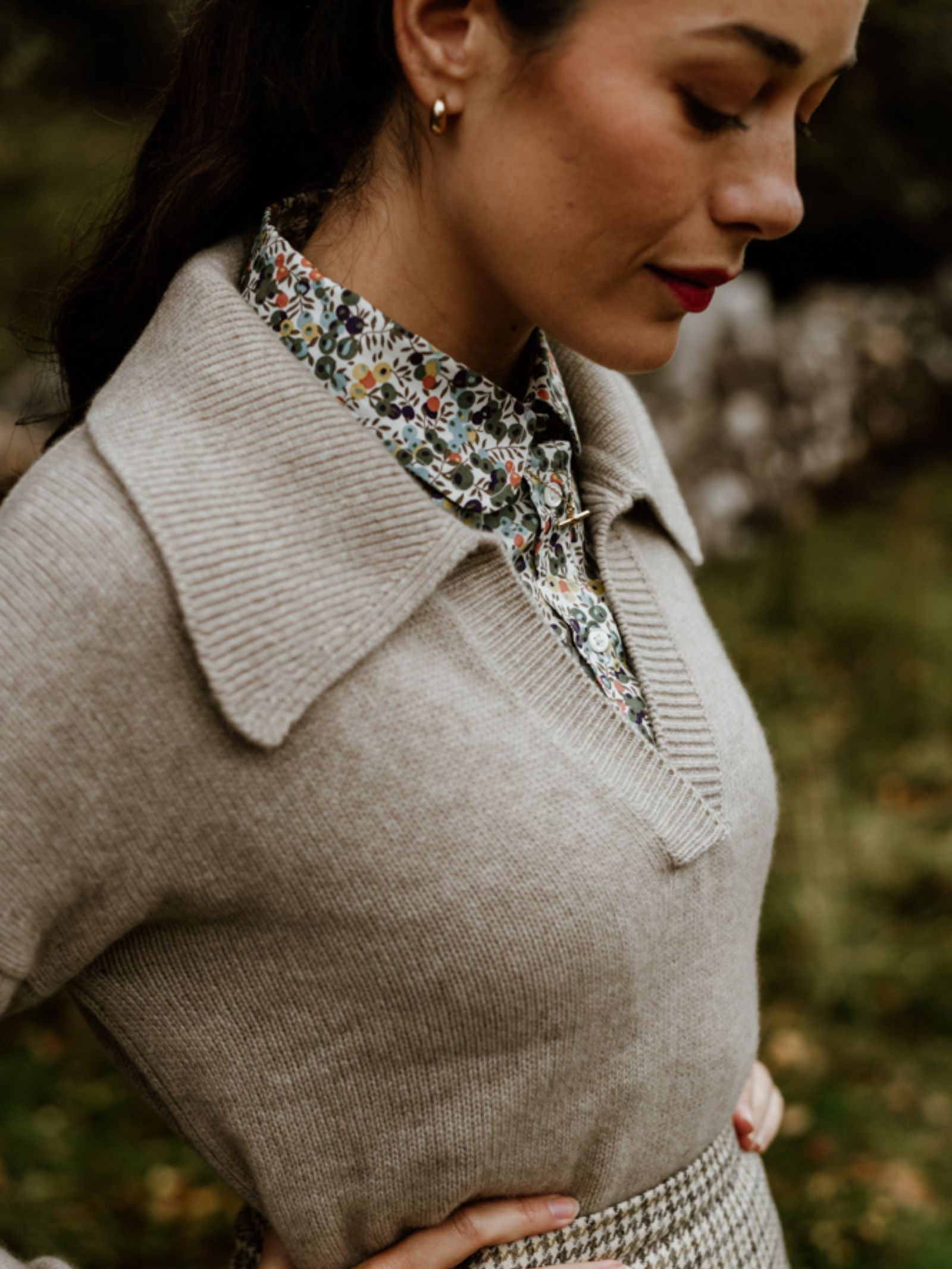 A woman stands outdoors in the Scottish Borders wearing Campbell's of Beauly's Auld Stock Cashmere Shirt Jumper, its wide collar layered over a floral shirt. Her hair is pulled back, gold hoops shine, and she gazes thoughtfully down, red lips striking.