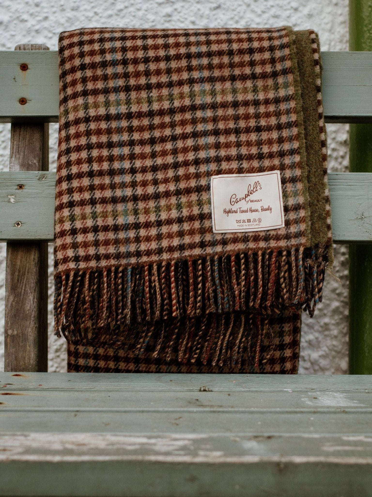 A Double-Faced Lambswool Throw from Campbell's of Beauly, in brown, red, and green tones with fringed edges, is draped over a weathered light blue bench. This cozy UK-made blanket features the Campbell's Highland Tweed House label.