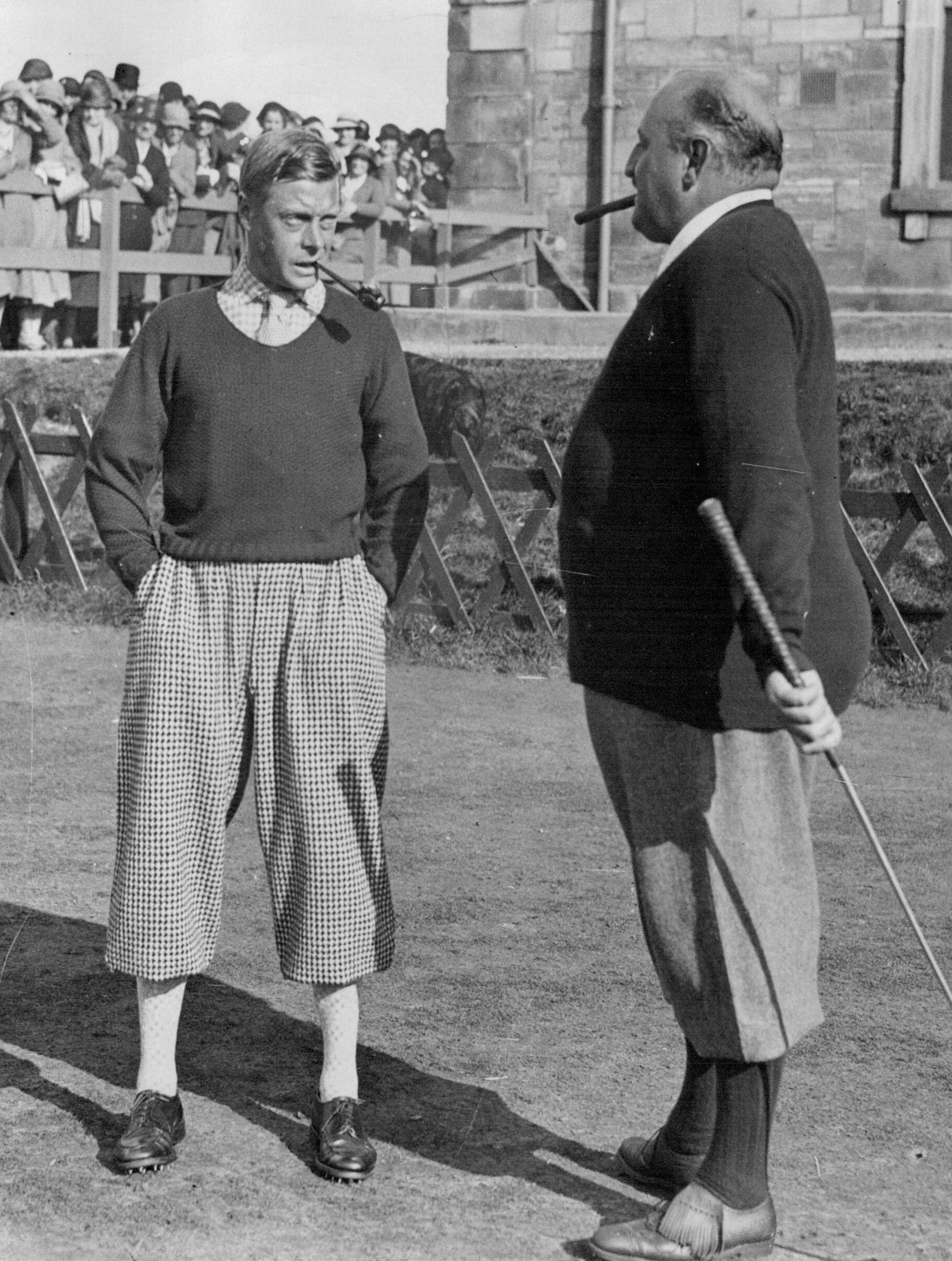 Two men in vintage golf attire stand outdoors, facing each other. Both are smoking cigars; one holds a golf club. A crowd and a stone building are visible in the background.