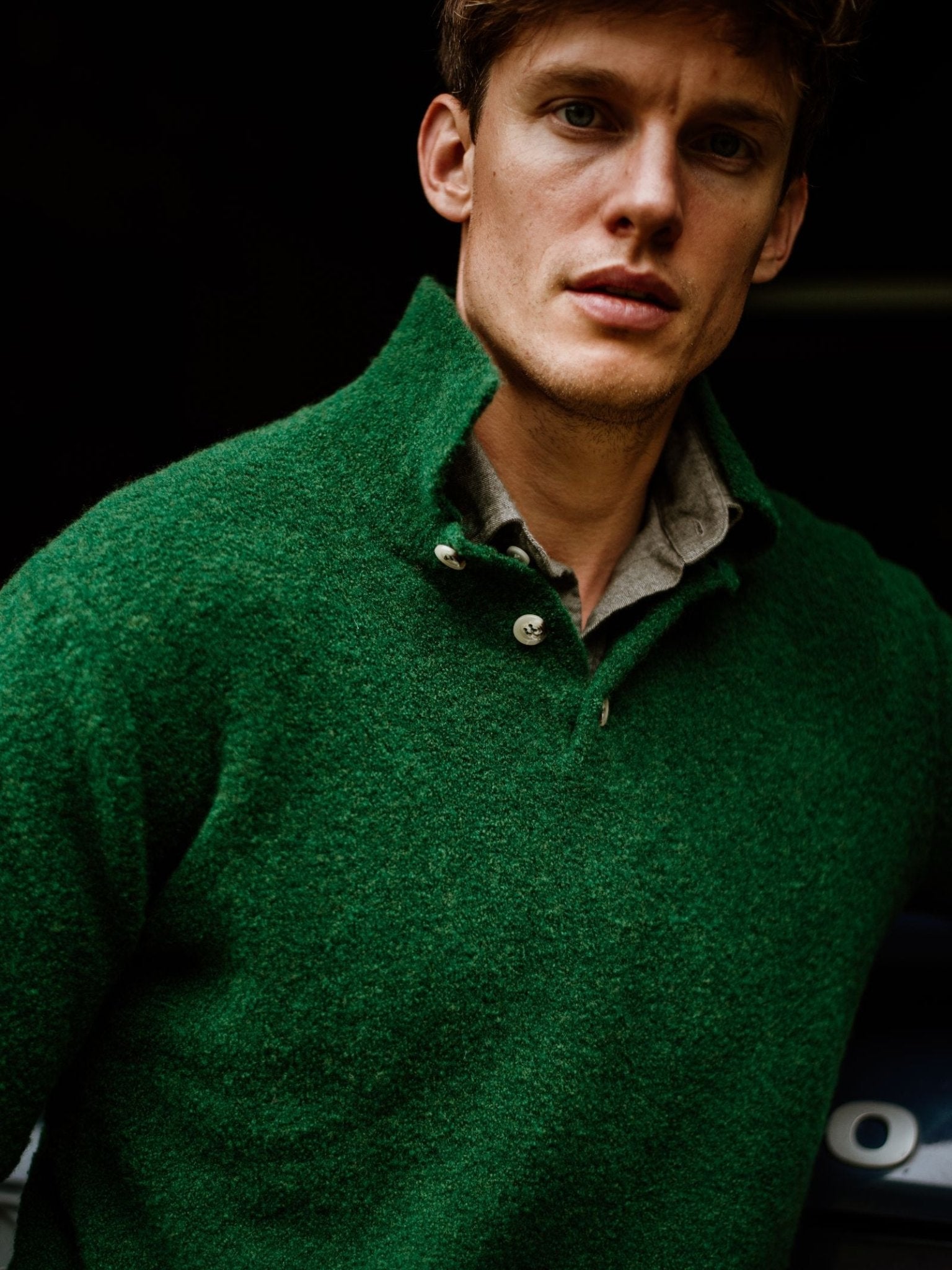 A man with short brown hair wears the Campbell's of Beauly Auld Stock Bouclé Neck Jumper in forest green over a light collared shirt, standing against a dark background.