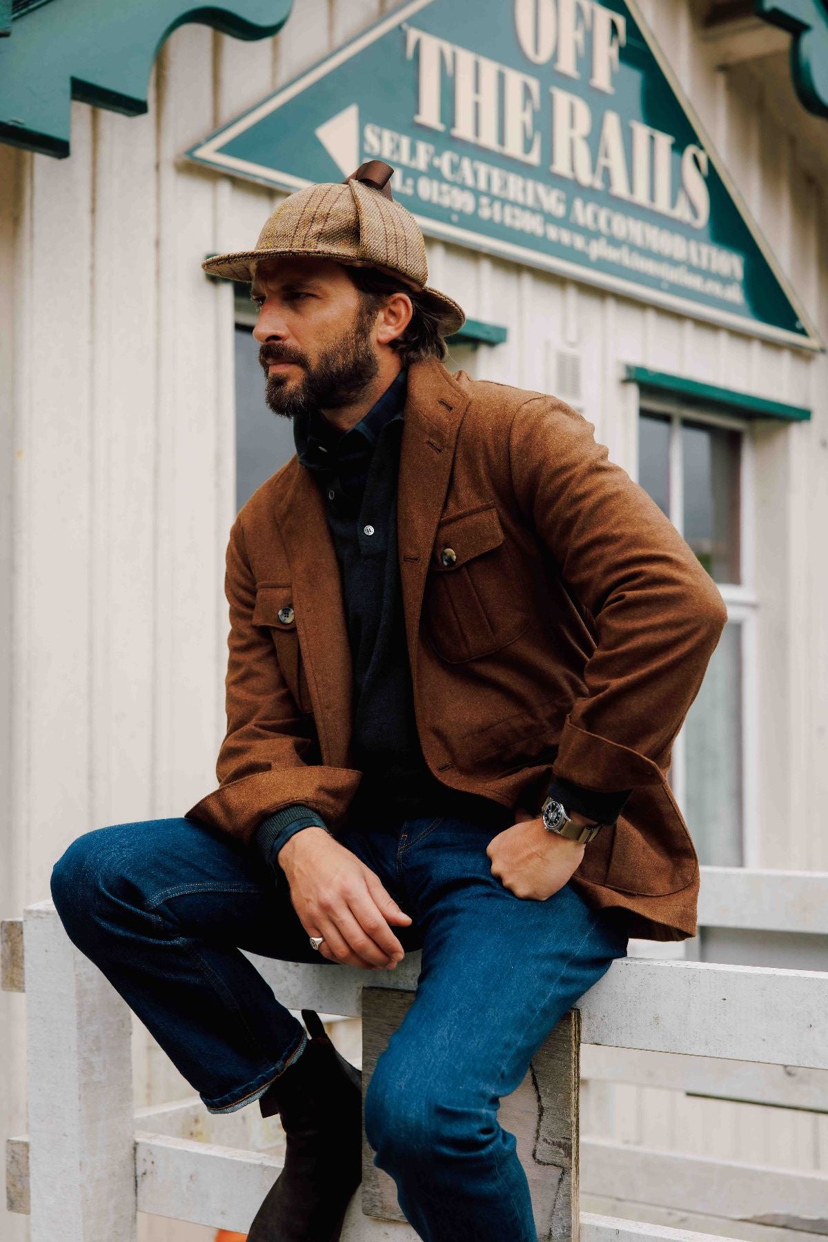 A bearded man wearing a brown jacket, dark shirt, blue jeans, and a plaid cap sits on a white wooden fence in front of a building with a green OFF THE RAILS sign.