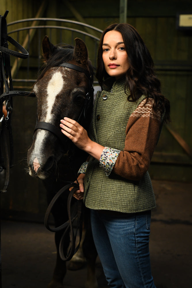 A woman with long dark hair wearing a green and brown jacket and blue jeans stands beside a black horse with a white stripe on its face, gently holding its bridle in a stable.