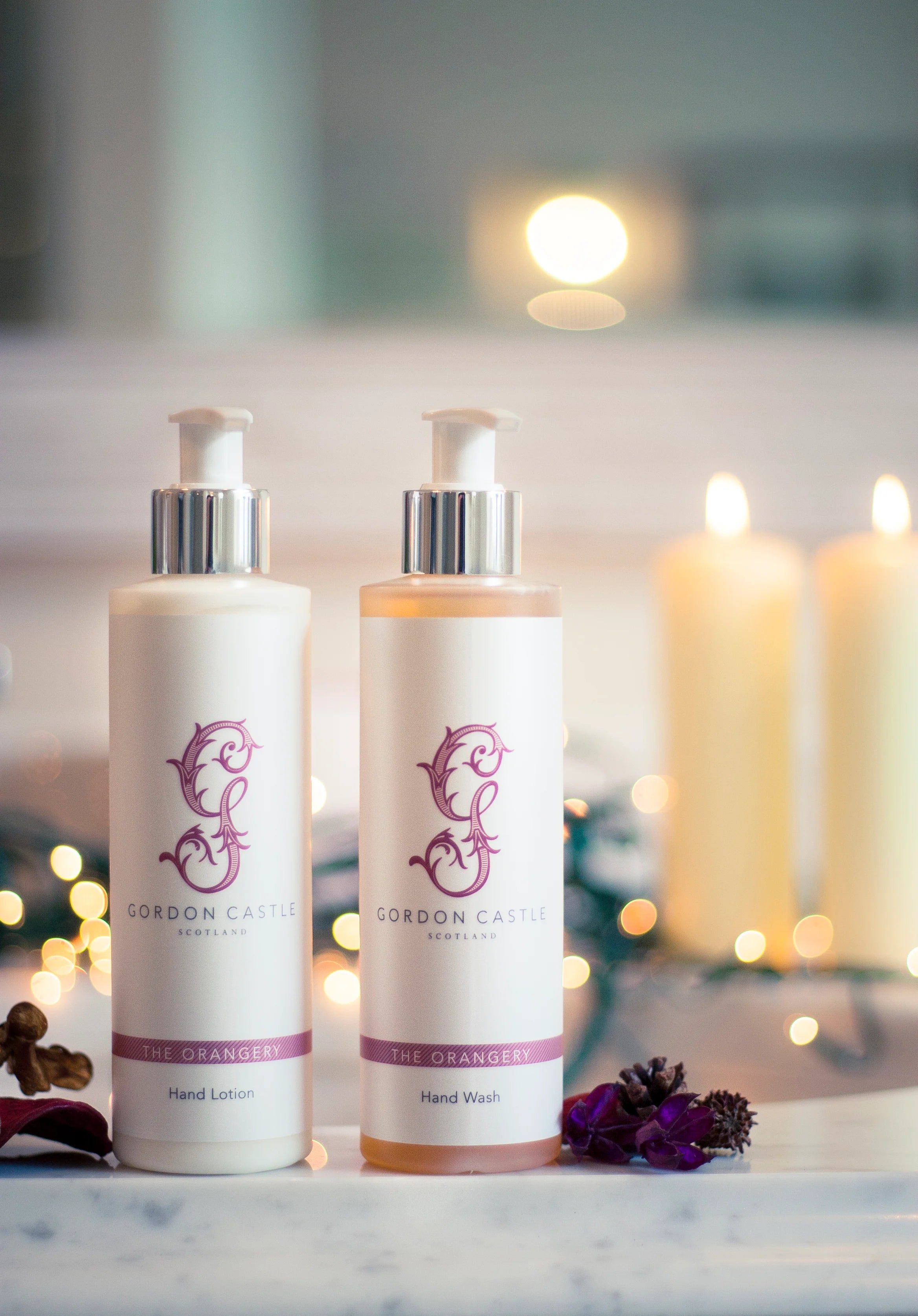 Two bottles, including Gordon Castle’s Orangery Hand Wash, sit on a counter. Candles and string lights glow in the background, with decorative greenery and a touch of citrus adding to the cozy, elegant scene.