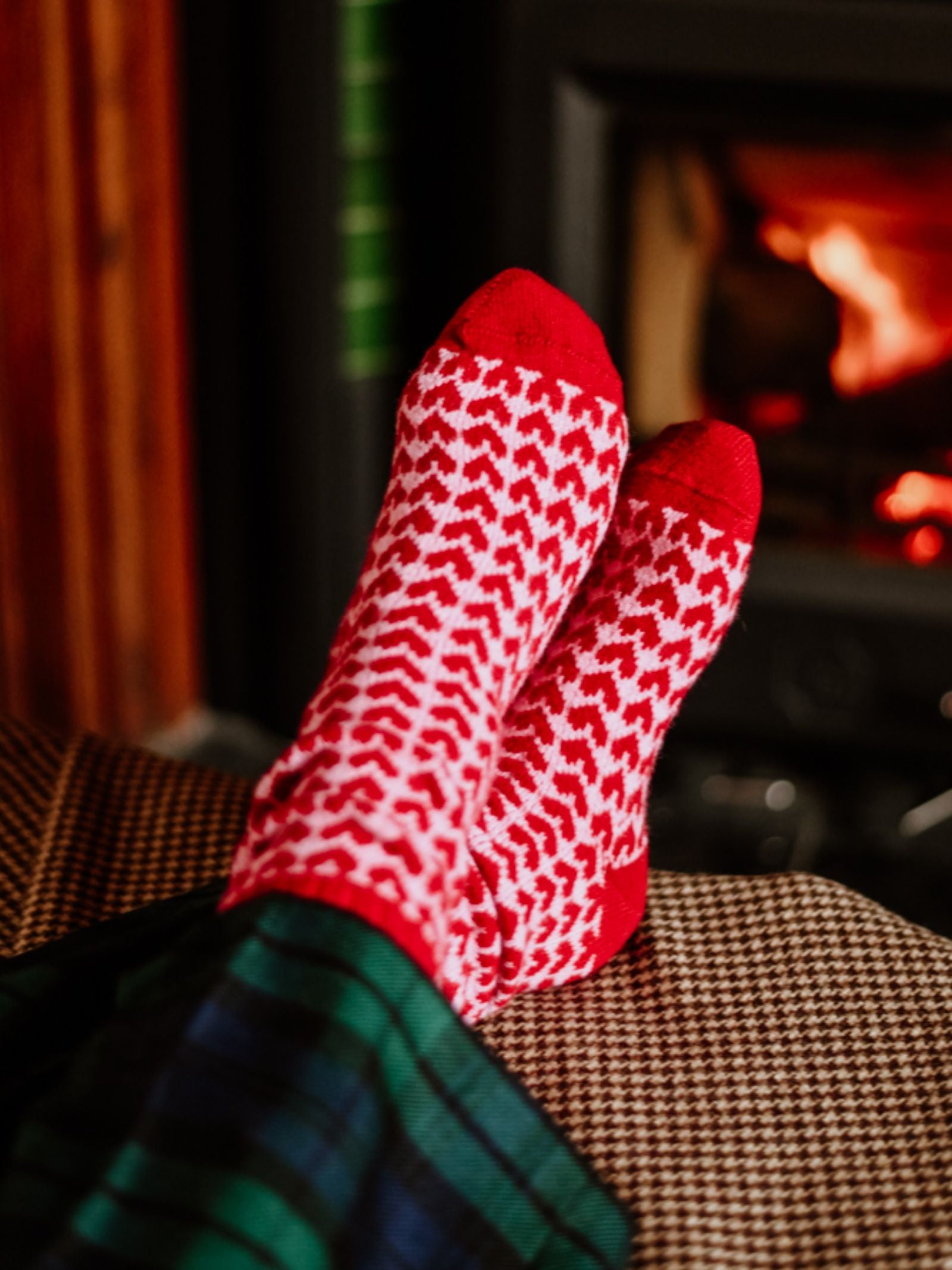 Cashmere Heart Socks by Campbell’s of Beauly in a red and white heart pattern are worn on relaxed feet, resting atop a brown houndstooth ottoman, while a warm fire glows in the background. The wearer pairs them with green and navy plaid pants.