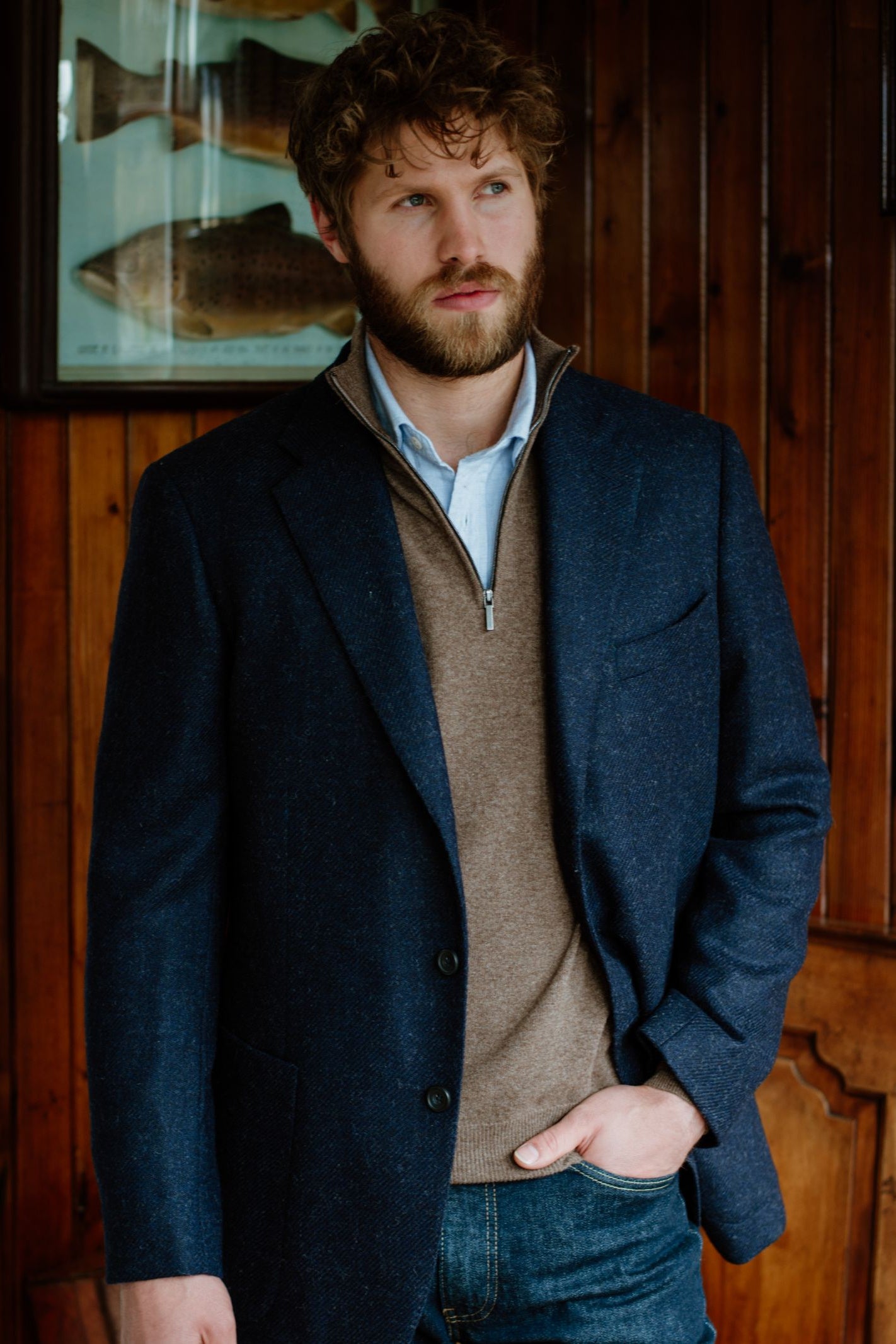 A man with wavy hair and a beard stands indoors against a wooden wall, wearing a Campbell's of Beauly Merino & Cashmere Quarter Zip Jumper over a light blue shirt and jeans. A framed fish illustration hangs behind him.