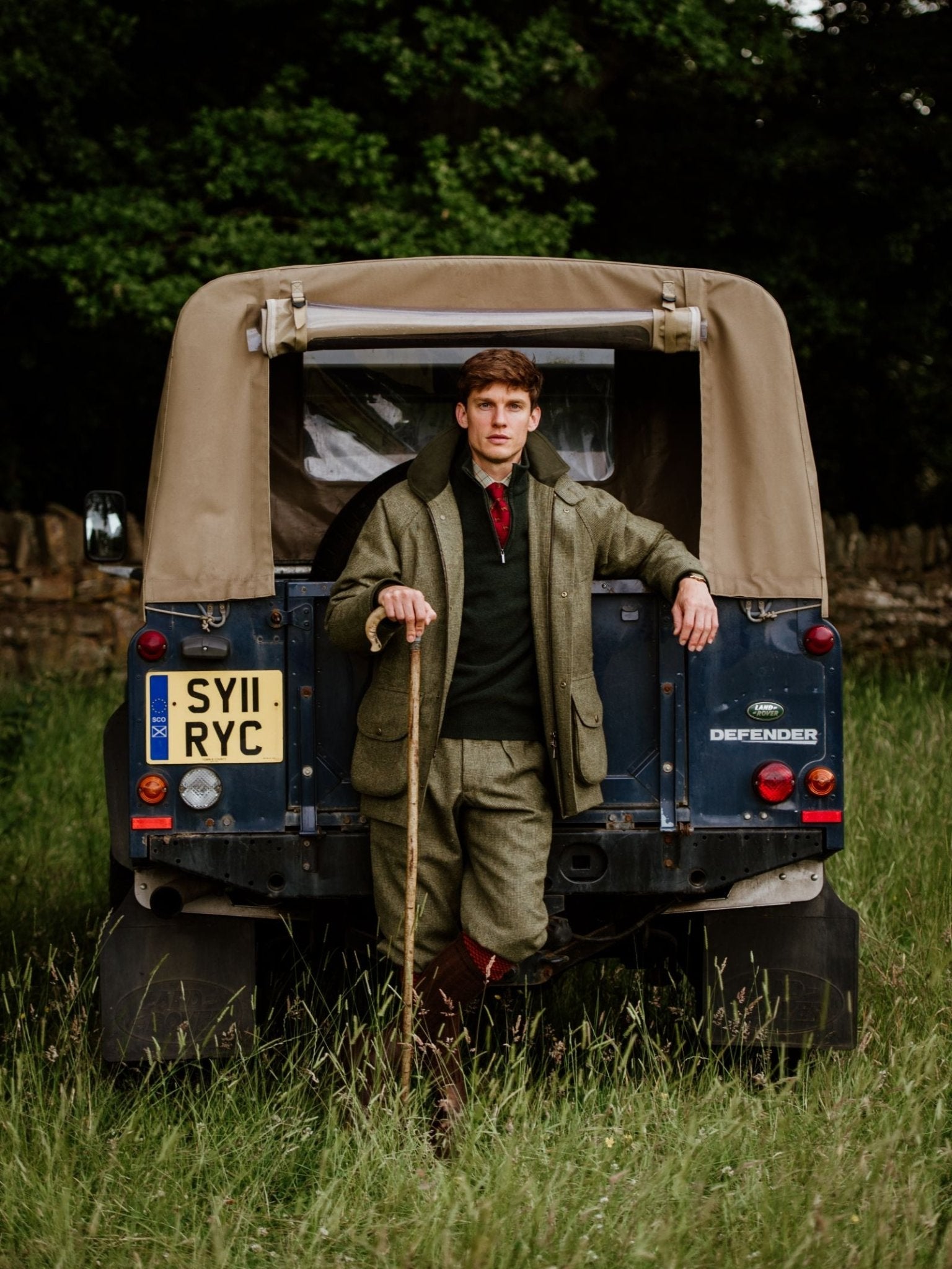 A man wearing Campbell's of Beauly Tweed Breeks and a flat cap stands in a grassy field, leaning against a Land Rover Defender. He holds a walking stick; trees and a stone wall are visible behind him.
