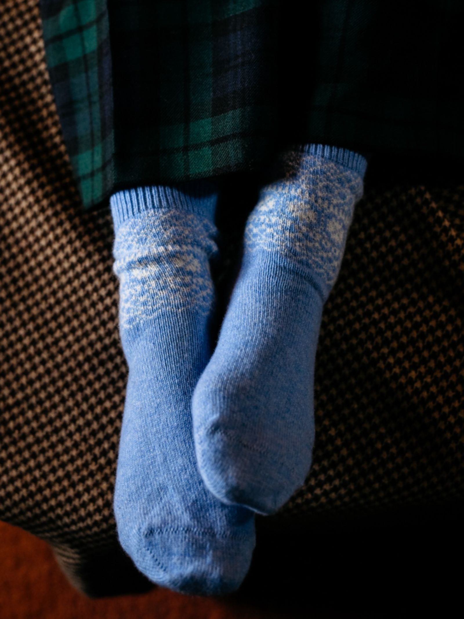 A person wears Campbell's of Beauly Two-Colour Fairisle Cashmere Socks in light blue with white patterns, sitting cross-ankled on a checkered surface and dressed in dark green and black plaid pants.