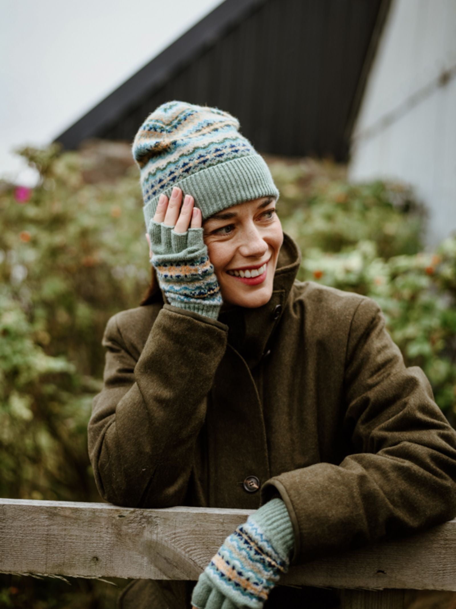 A person wears the Campbell's of Beauly Auld Stock Lambswool Fairisle Hat, standing by a wooden fence with greenery and a building behind—showcasing this cozy winter essential.