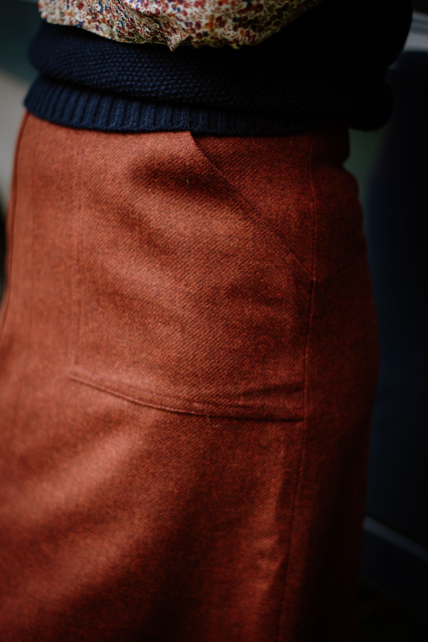 A close-up of a person wearing the Campbell's of Beauly Auld Stock A-Line Skirt in rust-colored British tweed with a front pocket, styled with a black knit sweater and a floral-patterned top.