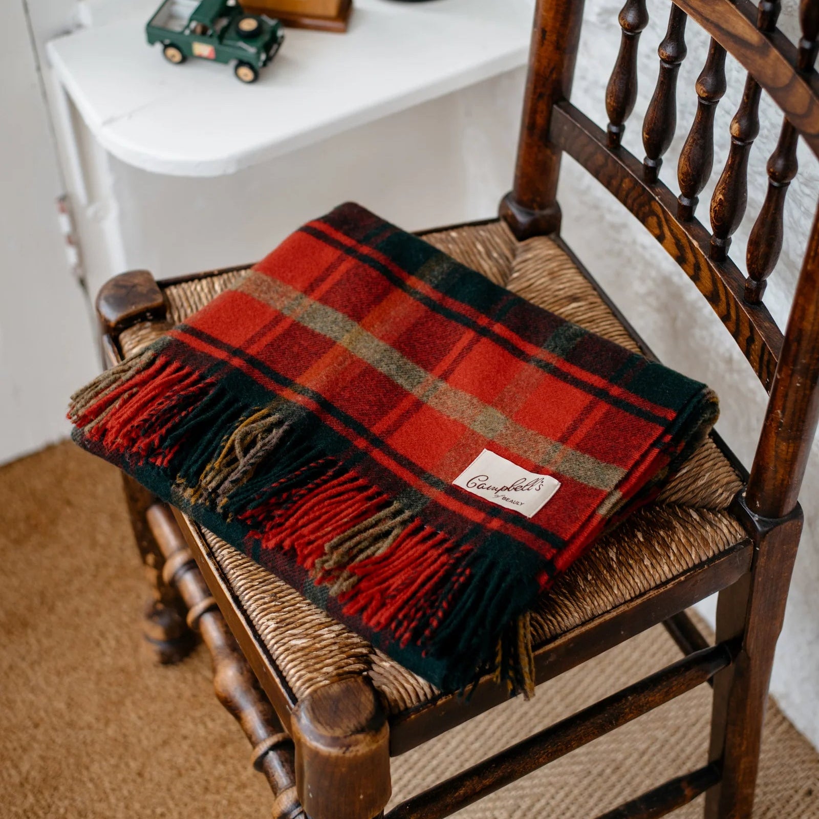 A red, green, and brown plaid wool scarf with fringed ends and a Campbell’s label is neatly folded on a wooden chair with a woven seat. A small green toy car sits on a white table in the background.