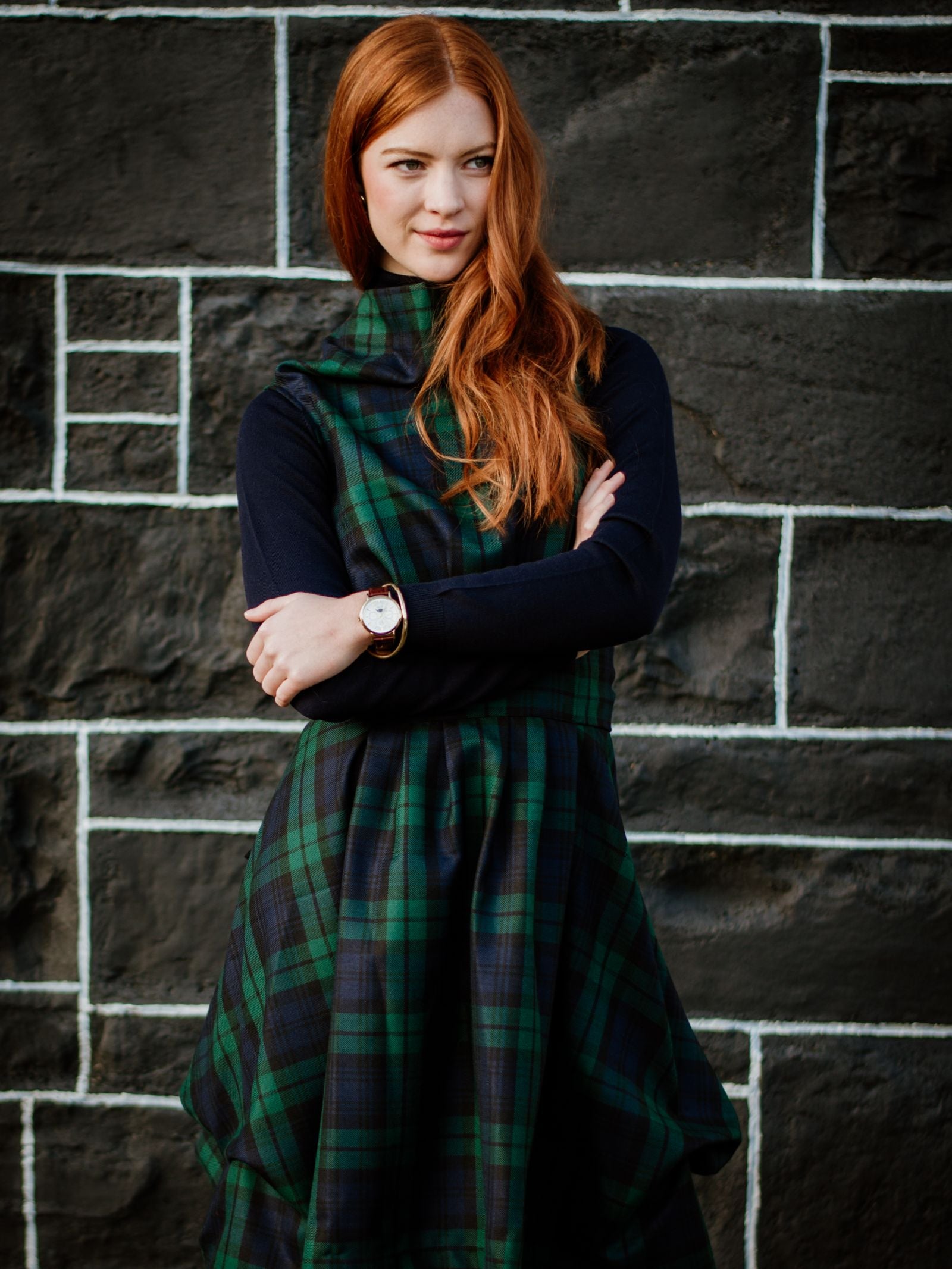 A woman with long red hair stands before a black stone wall, wearing a Campbell's of Beauly Reversible Wool Wrap Dress over a black top. Her arms are crossed as she looks to the side with a slight smile.