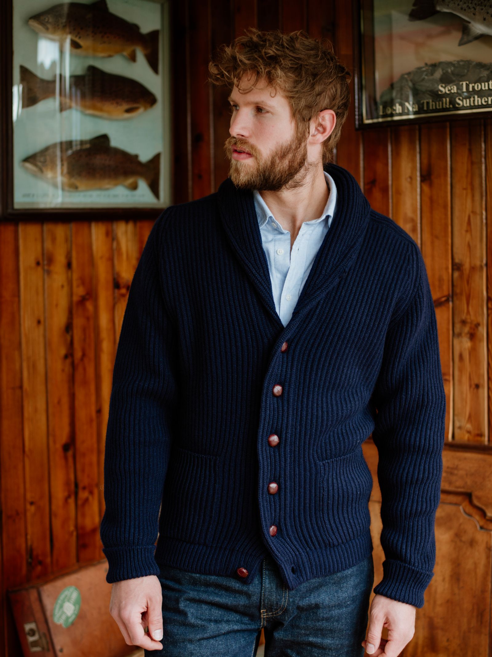 A man with curly hair and a beard wears the Campbell's of Beauly Cashmere Shawl Collar Cardigan in navy over a light blue shirt, standing indoors before wood-paneled walls with framed fish illustrations in the background.