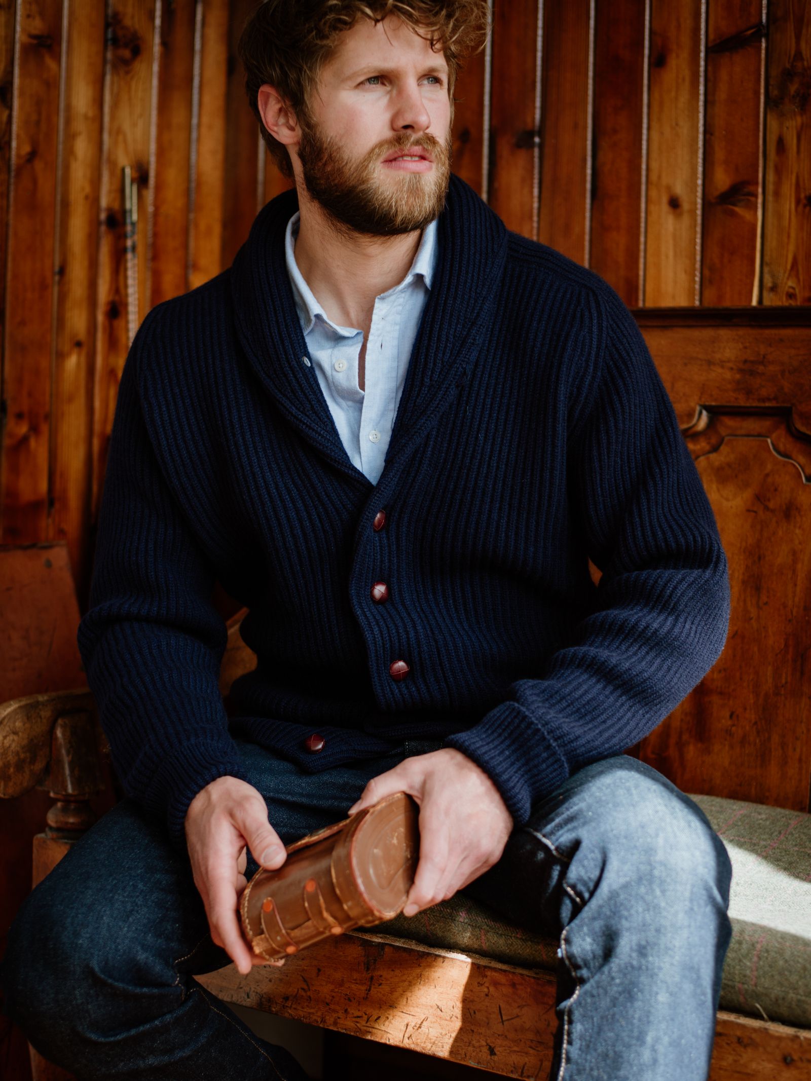 A bearded man in a Campbell's of Beauly Cashmere Shawl Collar Cardigan and jeans sits on a wooden bench, holding a brown ceramic mug as sunlight streams in from the side.